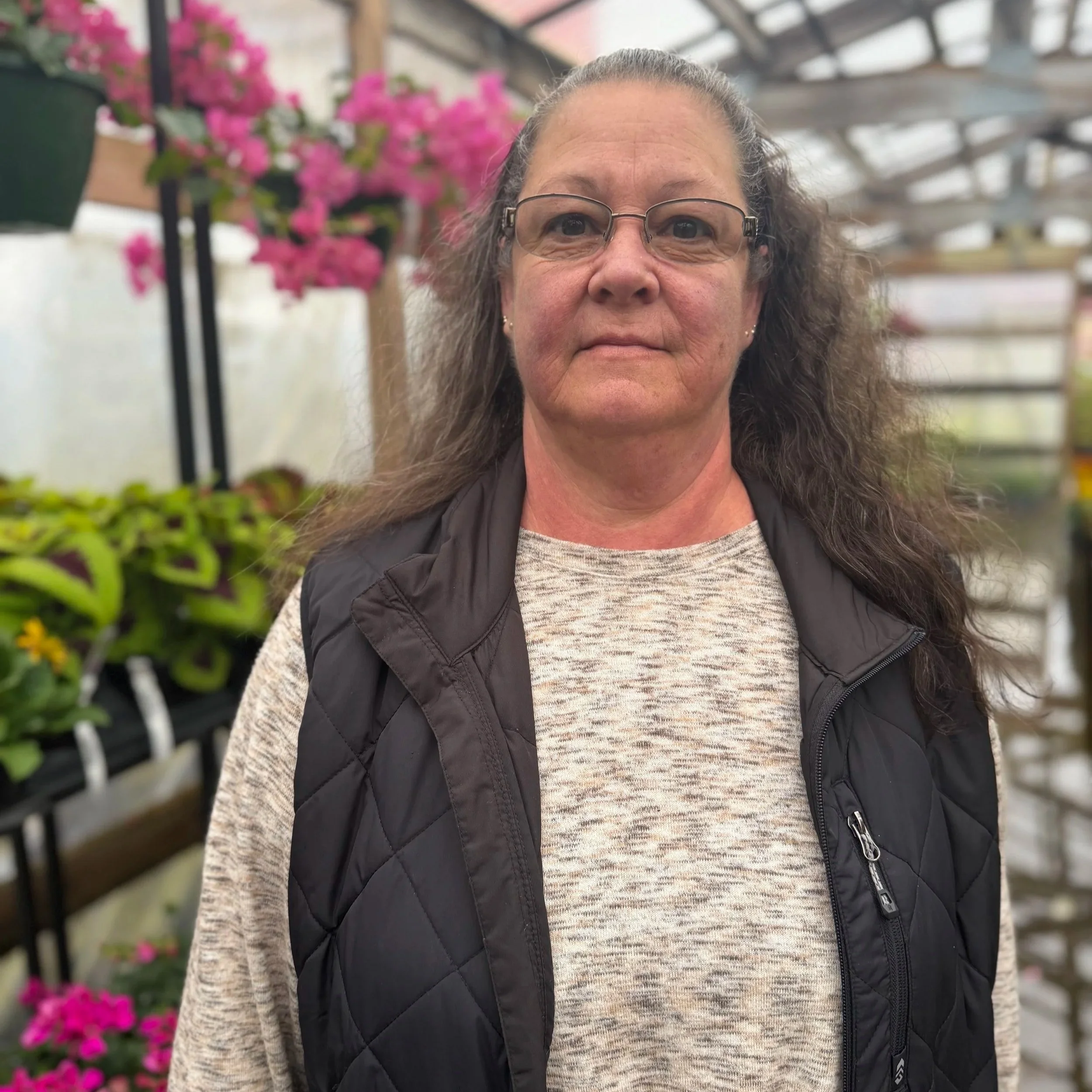 A woman with glasses, long gray hair, and wearing a beige shirt and black quilted vest standing inside a greenhouse surrounded by pink flowering plants and green foliage.