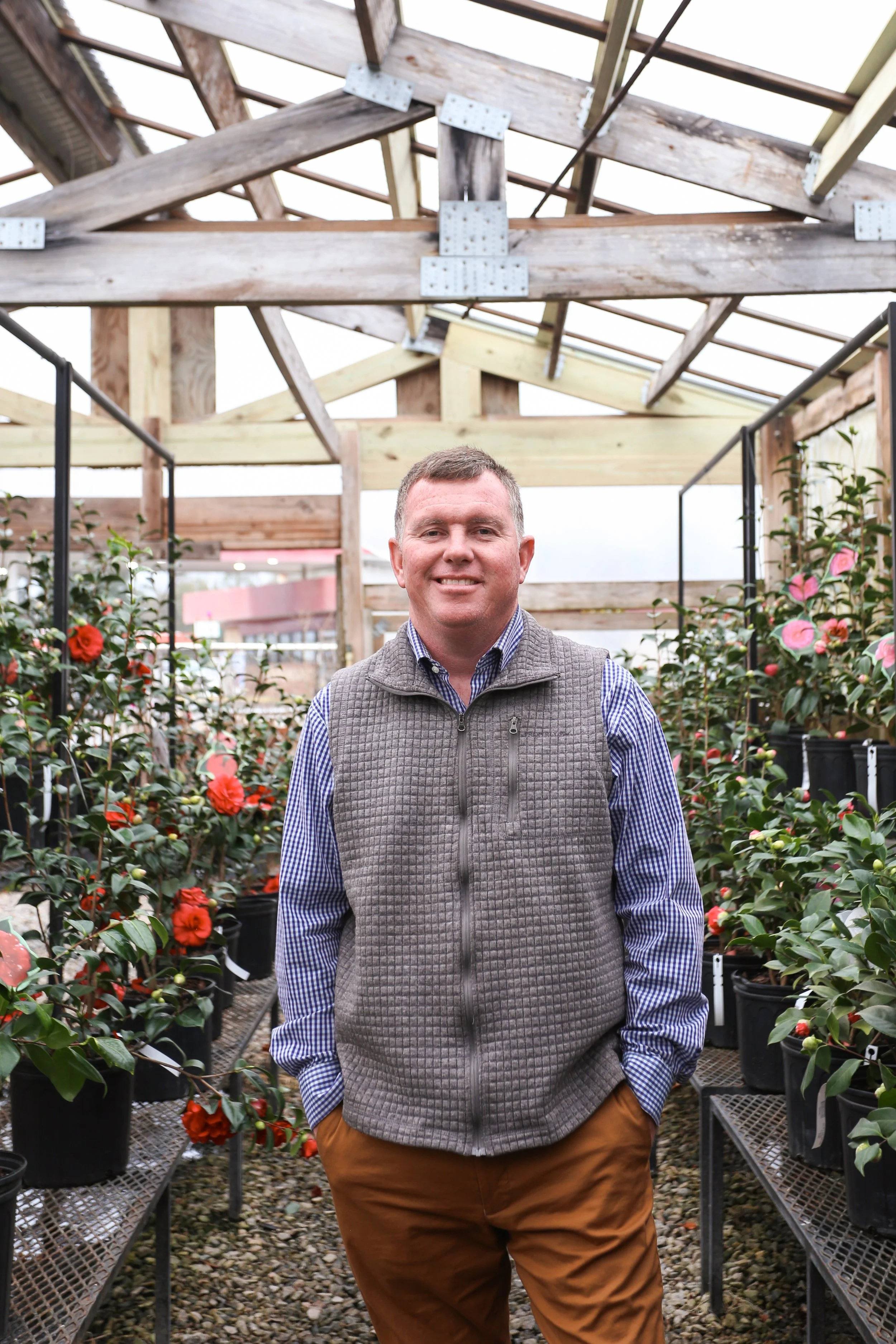 A man standing in a greenhouse surrounded by potted flowers with pink and red blooms, wearing a gray vest and brown pants.