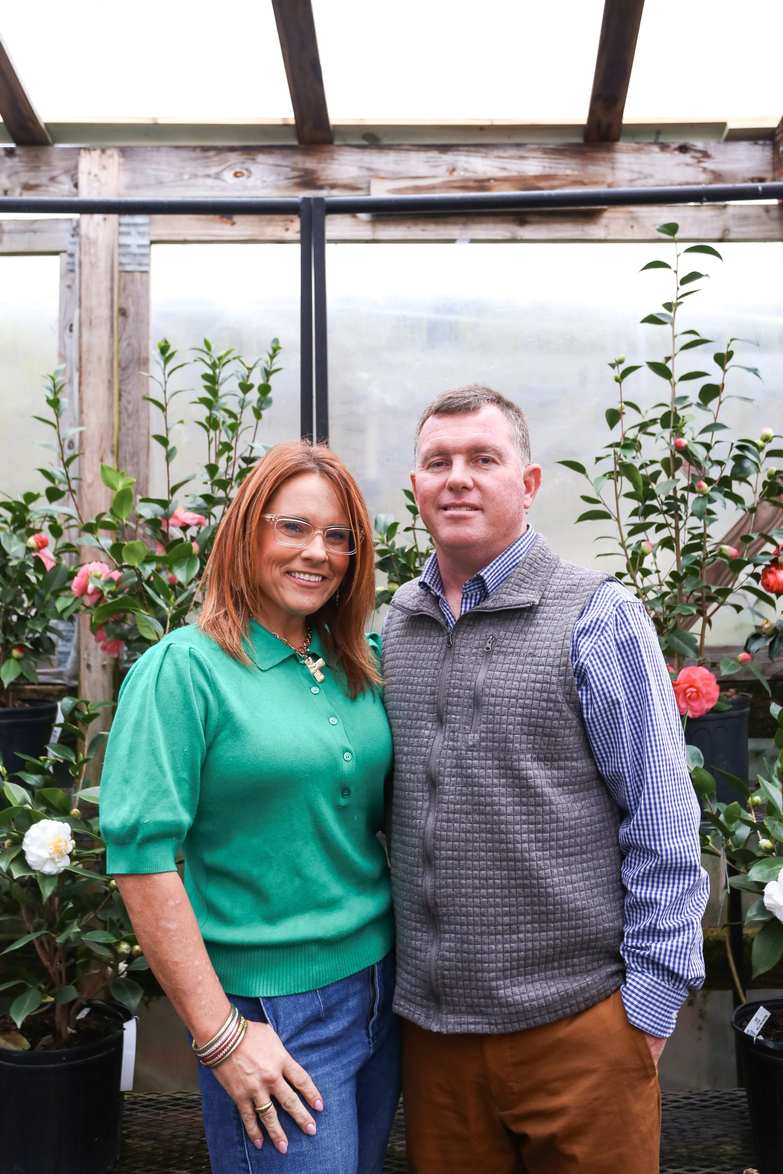 A woman and a man standing together inside a greenhouse, surrounded by potted camellia plants with pink and white flowers.