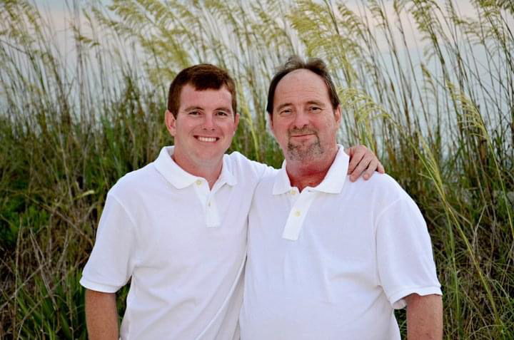 Two men standing outdoors in front of tall grass, both wearing white polo shirts, with one man having his arm around the other's shoulder, smiling at the camera.