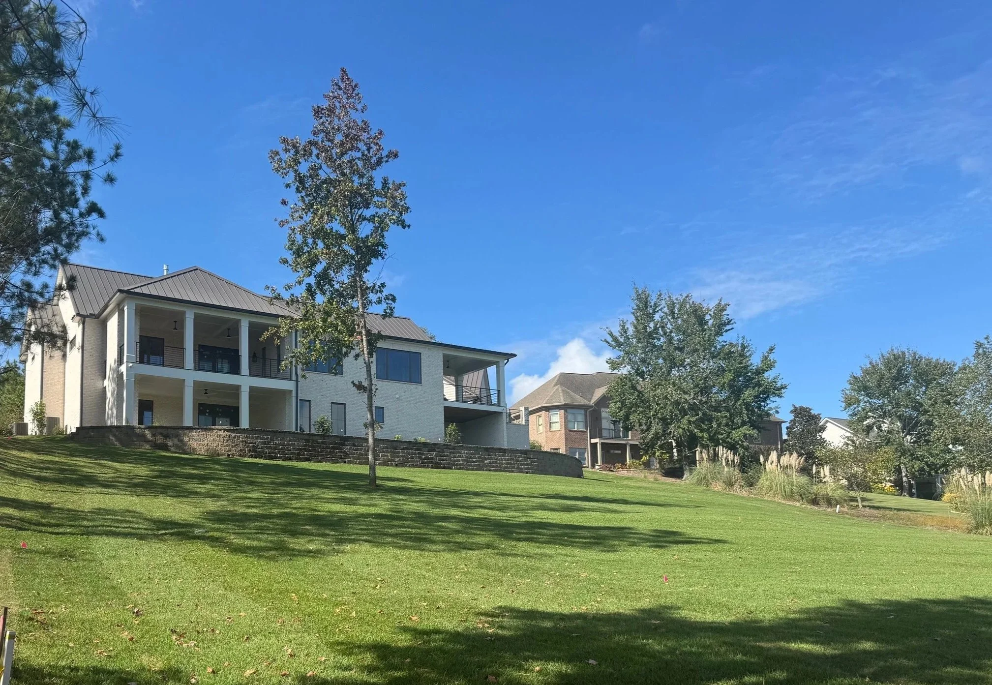 A modern multi-story house with white exterior walls, large windows, and balconies on a grassy hill, with trees and neighboring houses in the background under a bright blue sky.