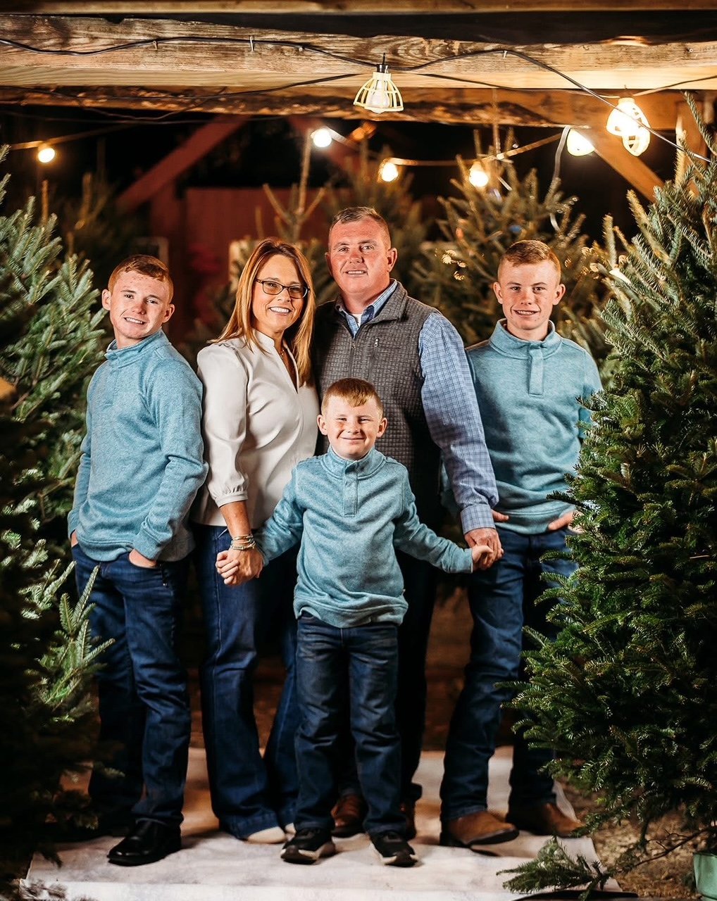 Family of five, including mother, father, and three boys, standing among Christmas trees in a rustic barn decorated with string lights at night.