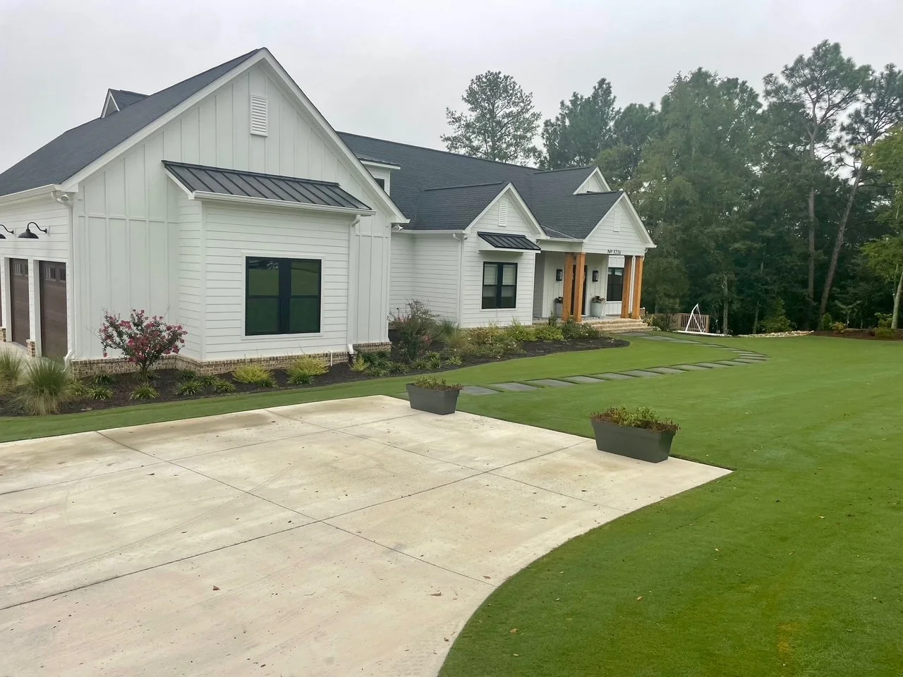 Modern white house with black roofing, large front yard with green lawn, and concrete driveway, surrounded by trees.
