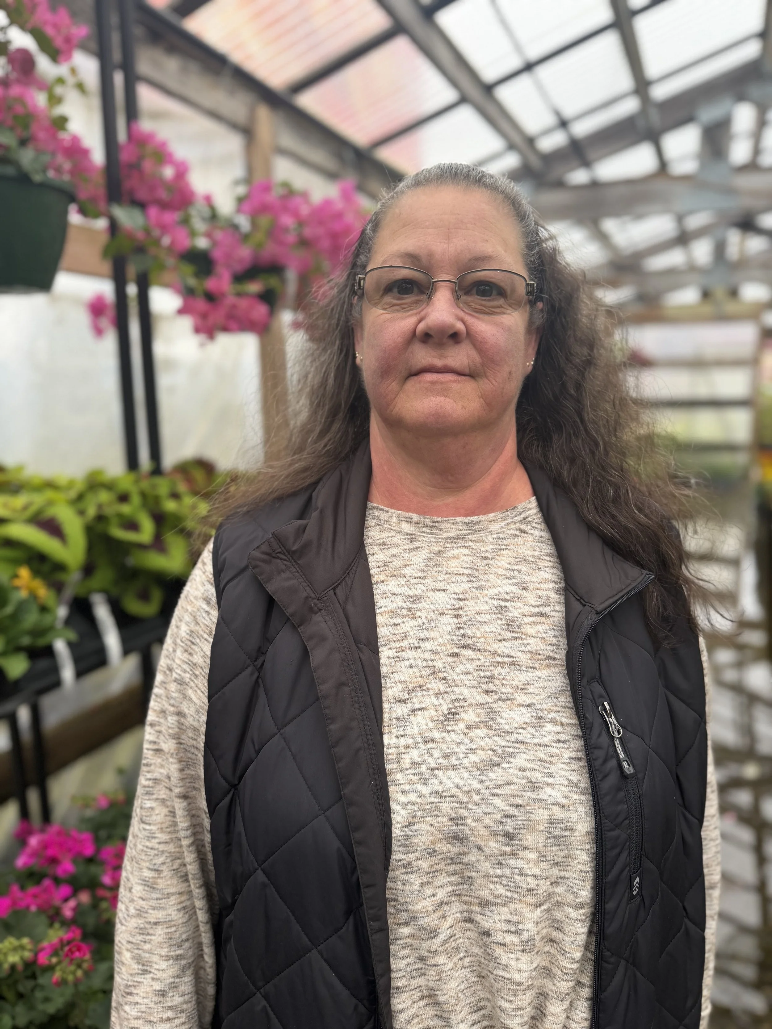 A woman with glasses, long gray hair, and wearing a beige shirt and black quilted vest standing inside a greenhouse surrounded by pink flowering plants and green foliage.
