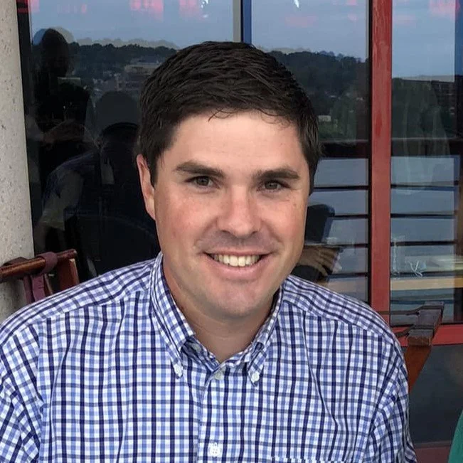A young man with dark hair smiling, wearing a plaid blue and white button-up shirt, sitting at an outdoor table, with a cityscape and water visible in the background.