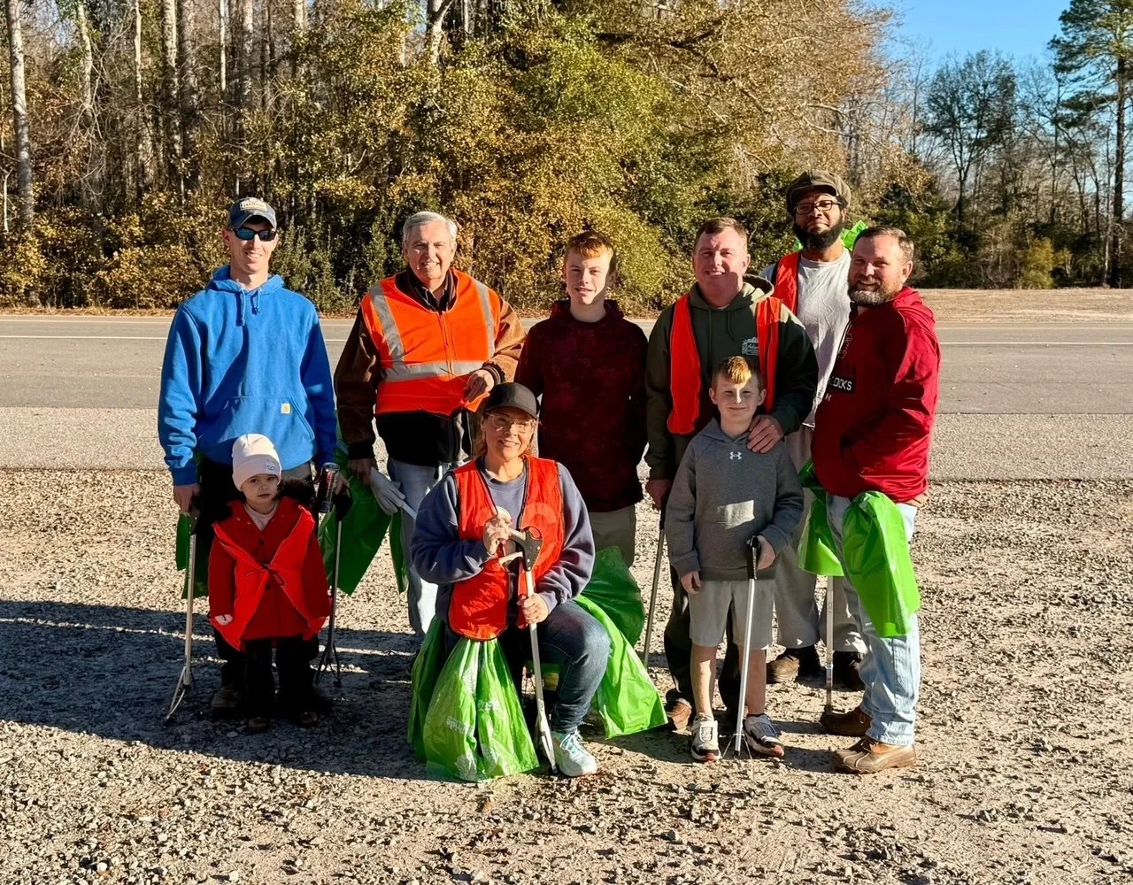 Group of people posing outdoors during a cleanup event, holding trash grabbers and wearing bright vests, with trees and an open road in the background.
