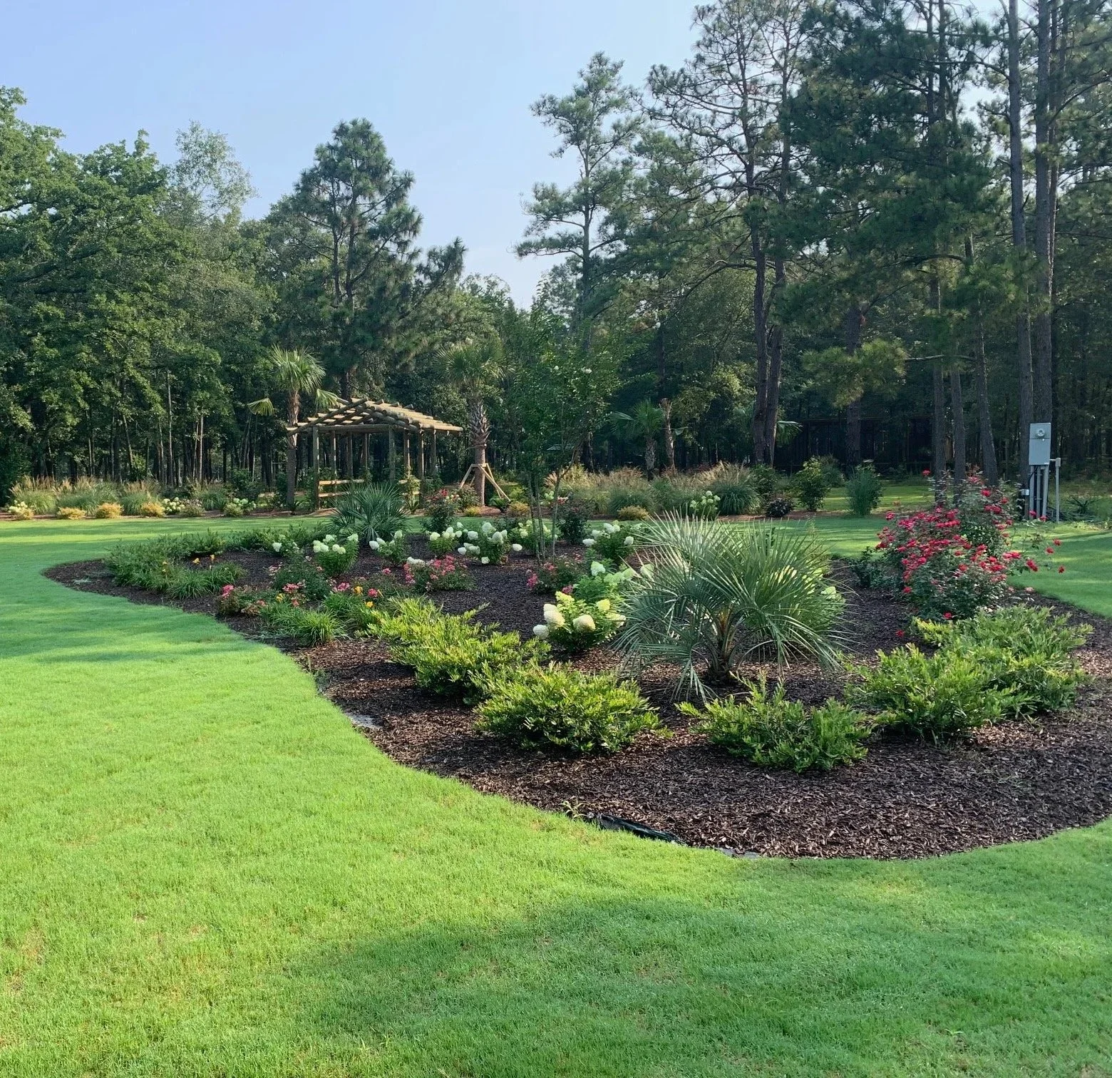 A landscaped garden with a flower bed containing various green plants and pink and white flowers, surrounded by a well-maintained grassy lawn and trees in the background.