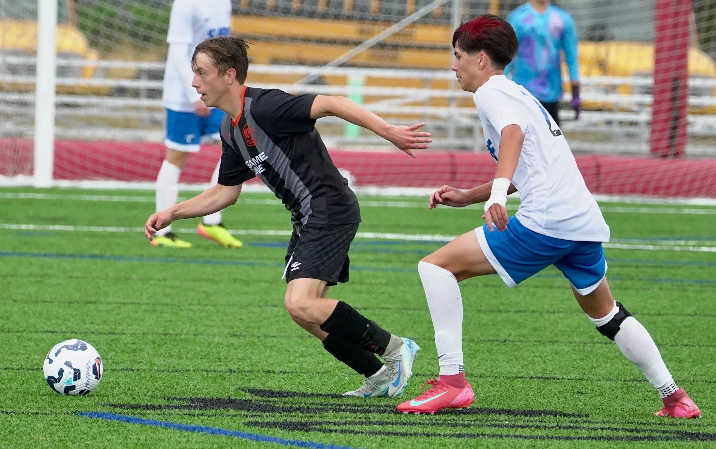 Two young male soccer players chase after a soccer ball on a field as a goalkeeper stands behind them. One player is wearing a black uniform and the other a white and blue uniform.