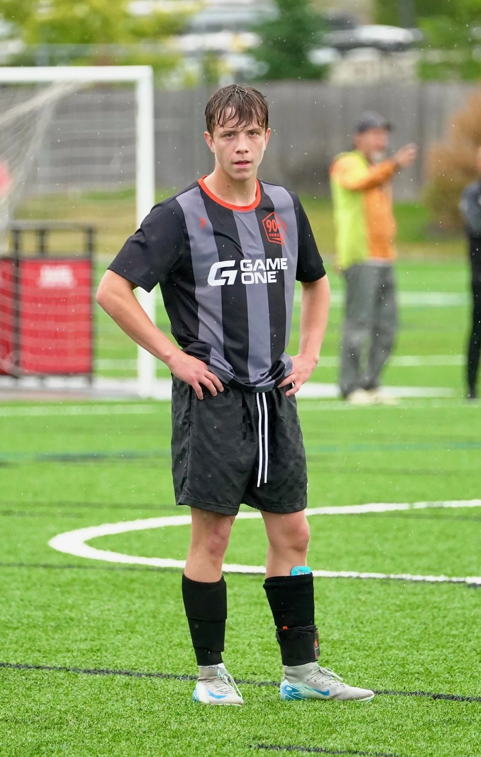 A young male soccer player standing on a wet field wearing a black and gray uniform with shorts, socks, and cleats, with hands on hips and wet hair, during a rainy day.