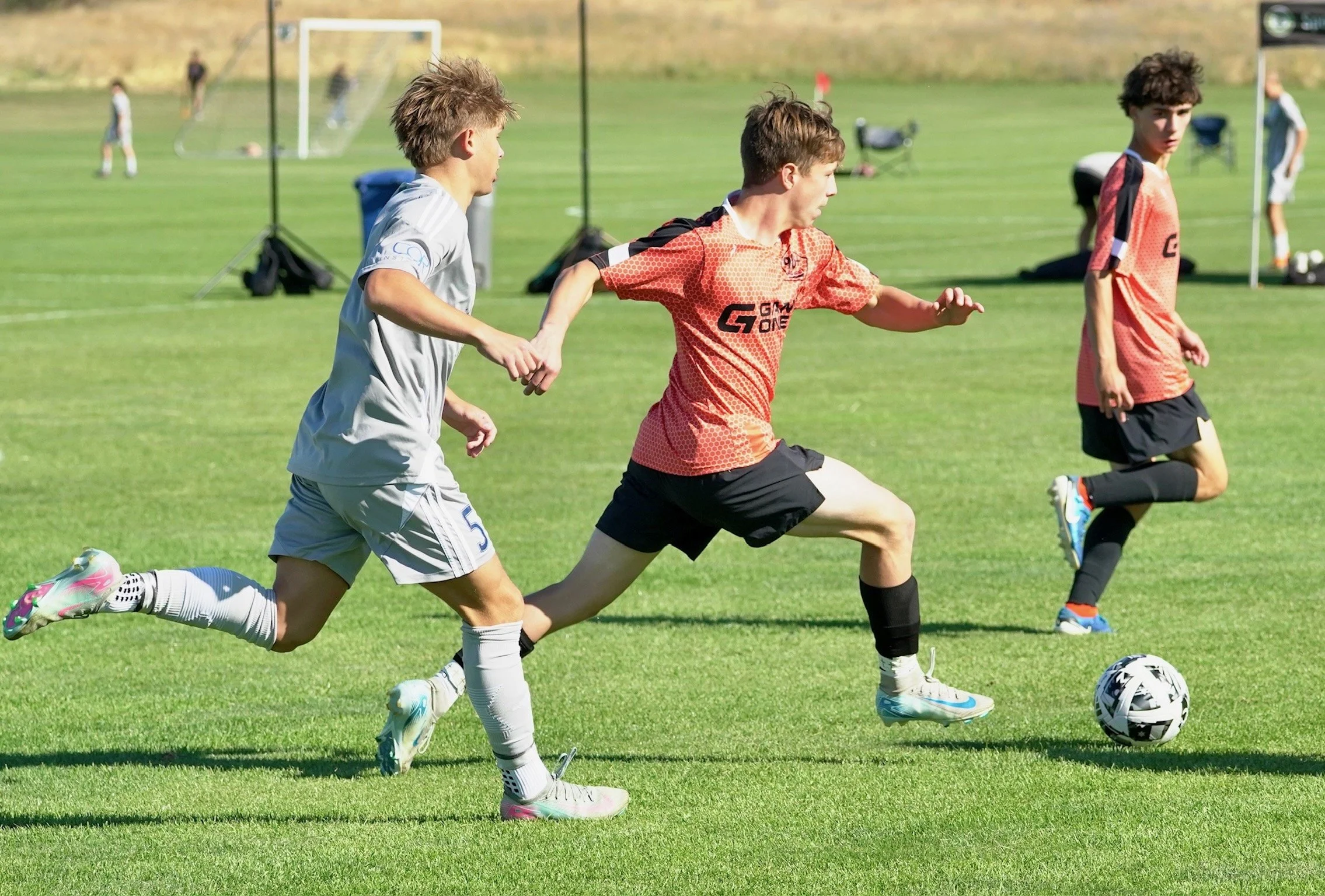 Three young boys playing soccer on a green field during daytime, with a goalpost and other players in the background.
