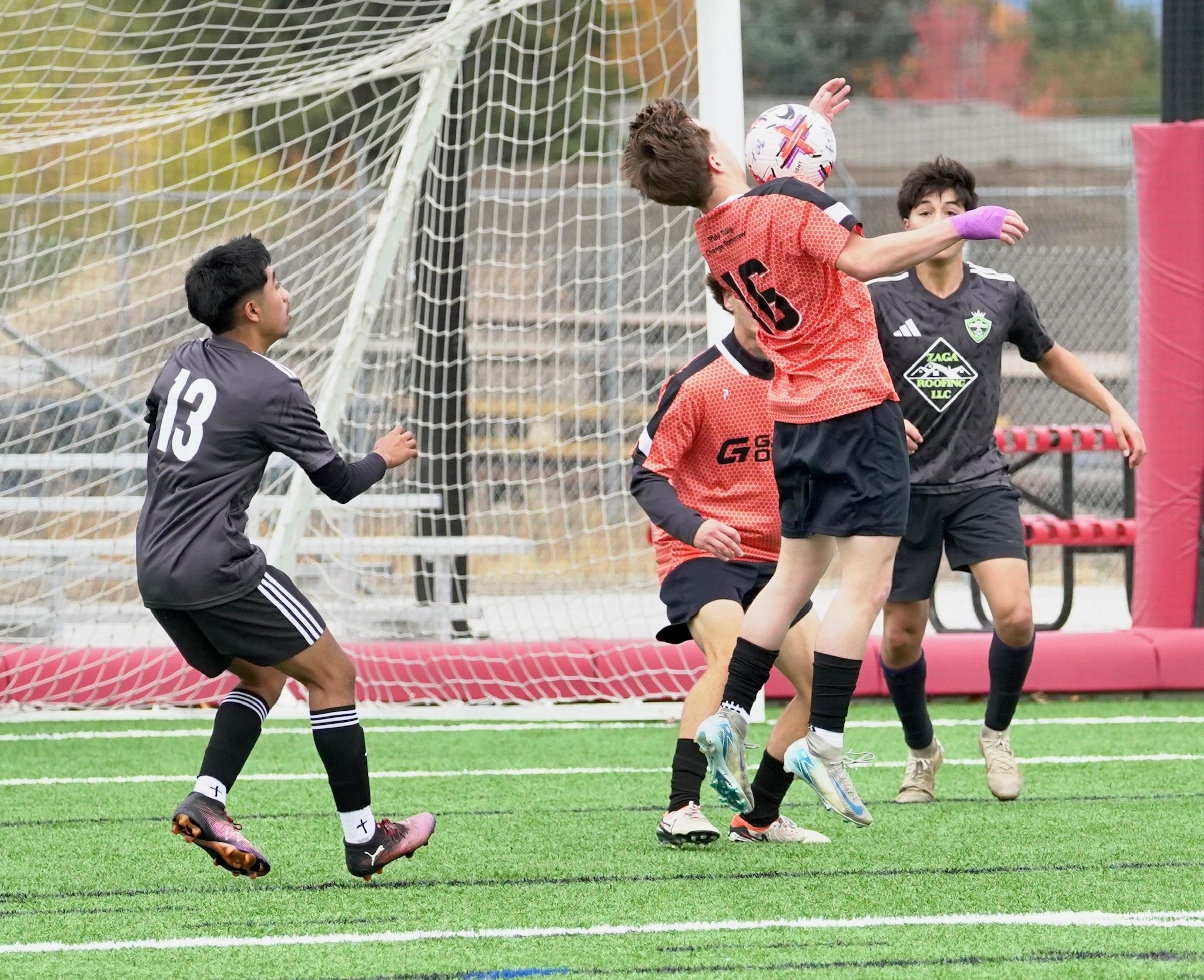 Young soccer players in black and red jerseys jumping and preparing to head the ball near the goal, with a goalkeeper observing nearby on a green turf field.