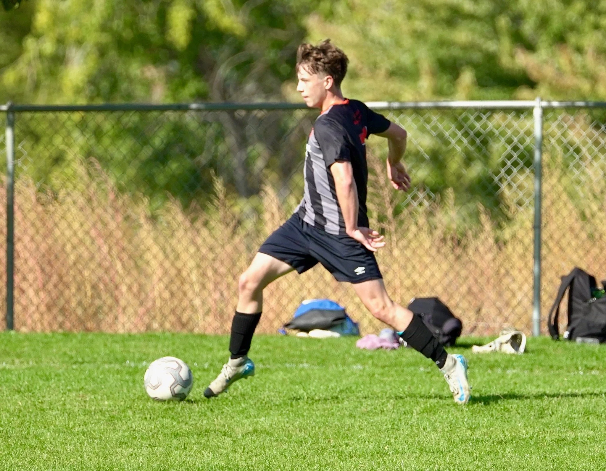 A young male soccer player in black uniform is kicking a soccer ball on a green field with a chain-link fence and trees in the background.