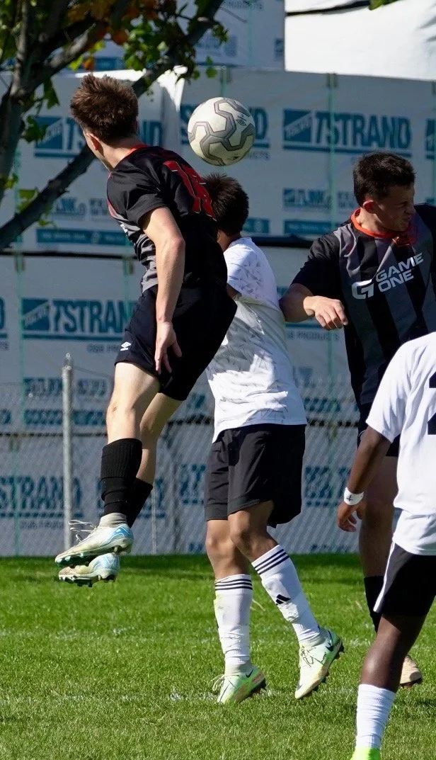 Two young male soccer players competing for a ball in mid-air during a game on a grassy field, with a few other players visible and a background of fencing and advertising banners.