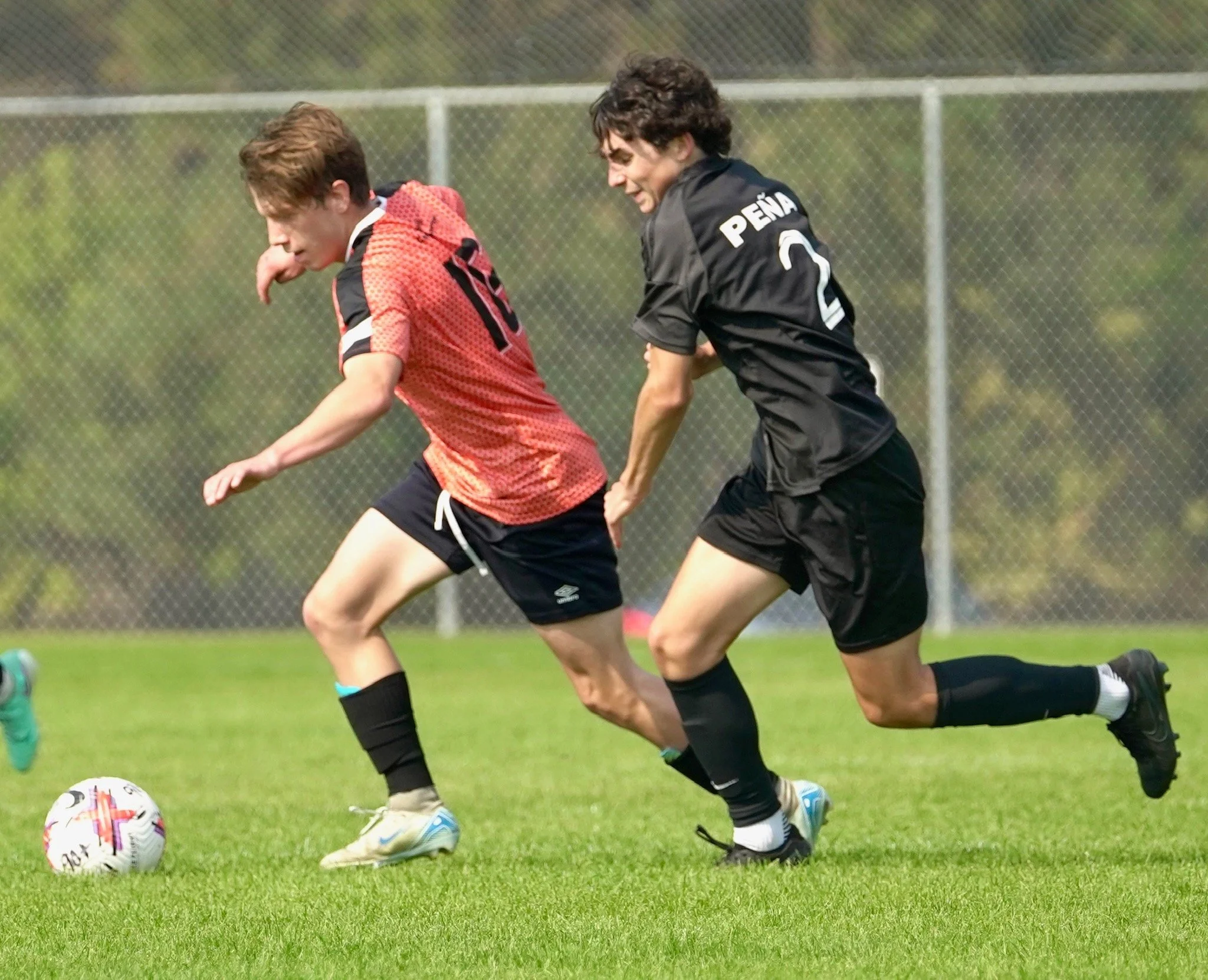 Two young male soccer players compete for the ball on a grassy field, with one in a red jersey and the other in a black jersey.