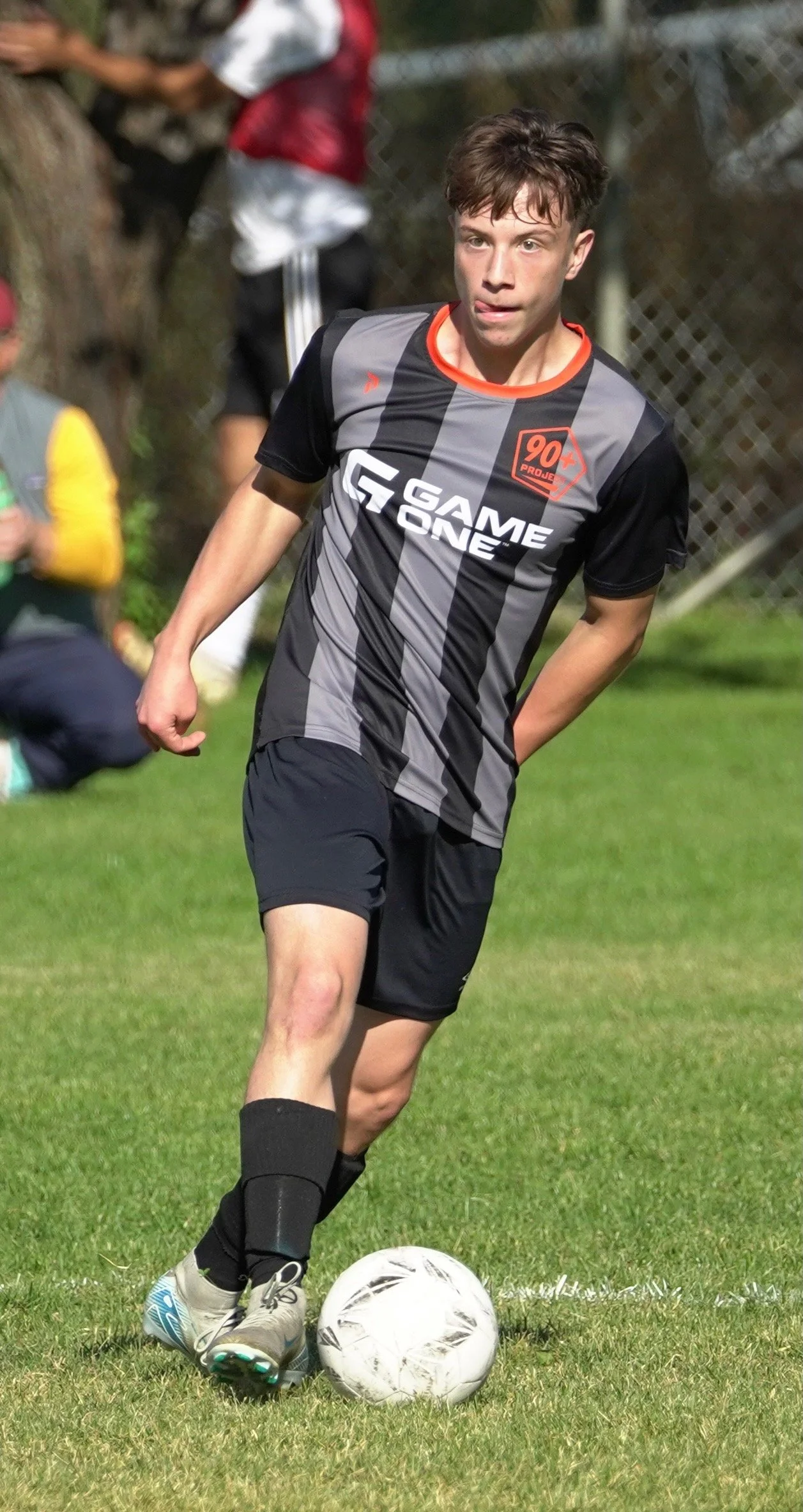 A young man playing soccer, about to kick a white soccer ball on a grassy field. He is wearing a black and gray jersey with a red collar, black shorts, black socks, and gray soccer cleats. In the background, there are people and a chain-link fence.