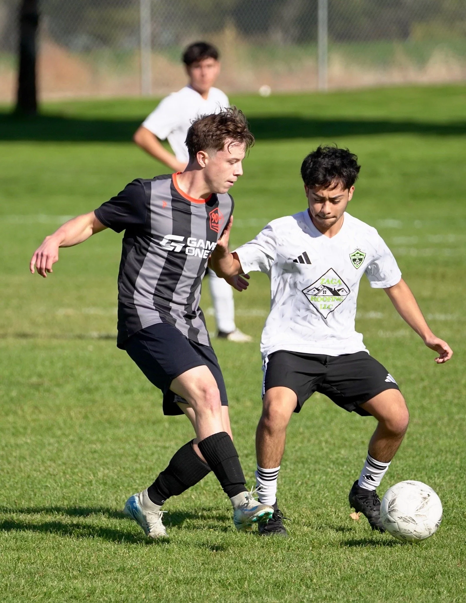 Two soccer players competing for the ball on a grassy field, with a third player in the background.
