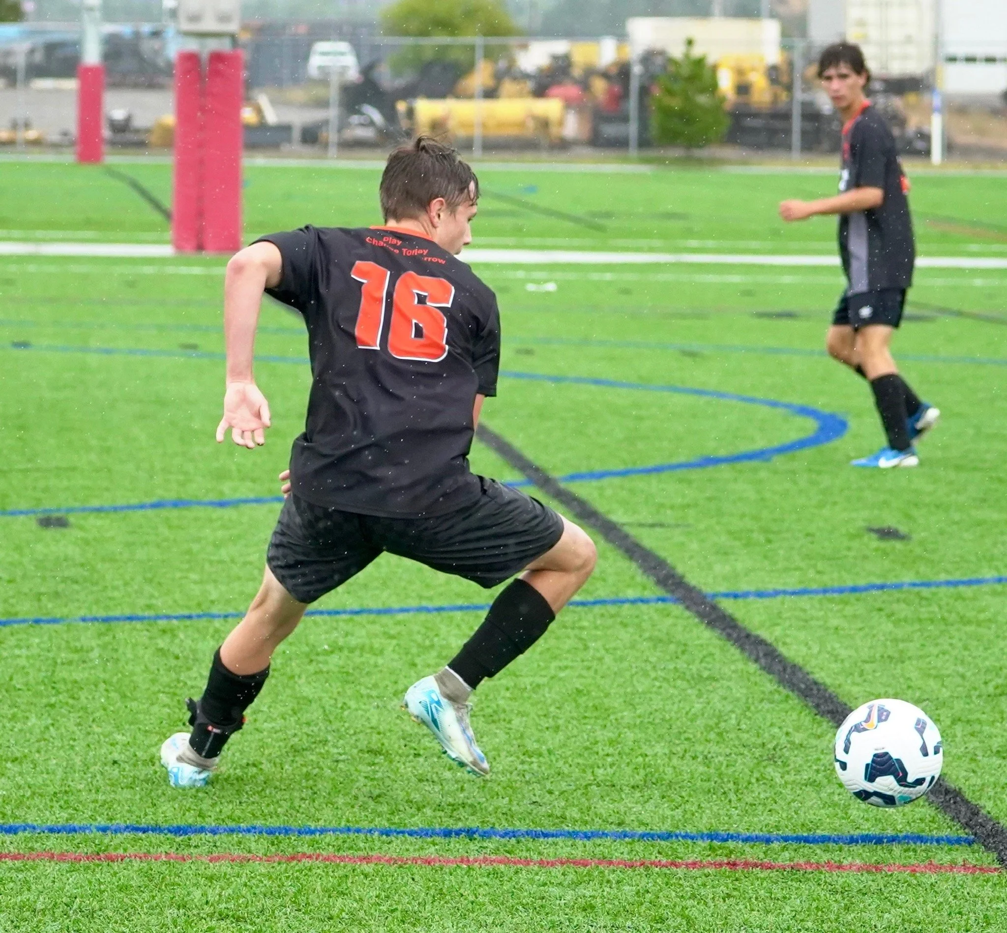 Young soccer player in black uniform dribbling a soccer ball on a rainy field, with another player in the background.