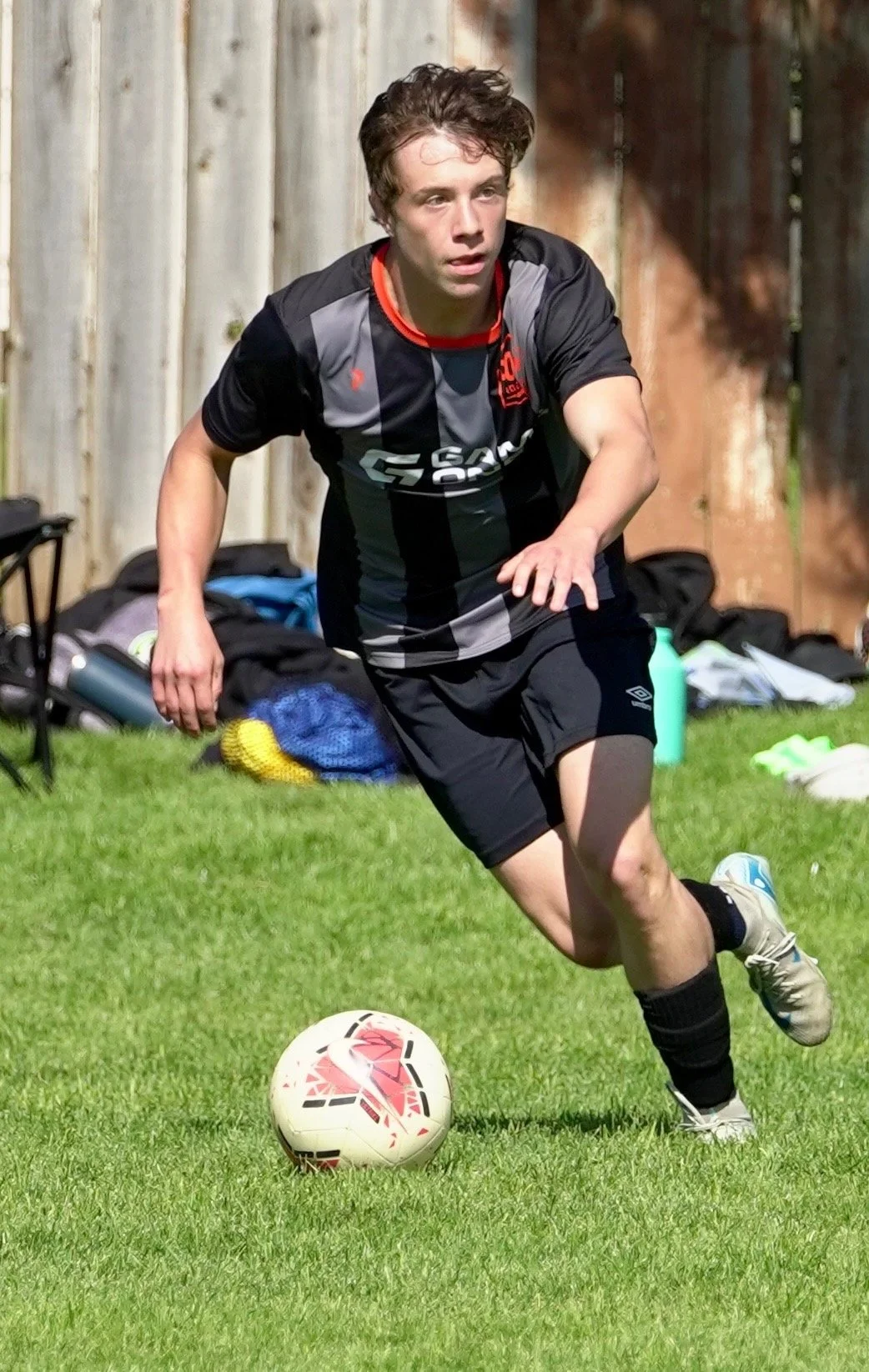 Young man playing soccer outdoors on a grassy field, wearing a black and gray sports jersey and shorts, with a soccer ball in front of him.