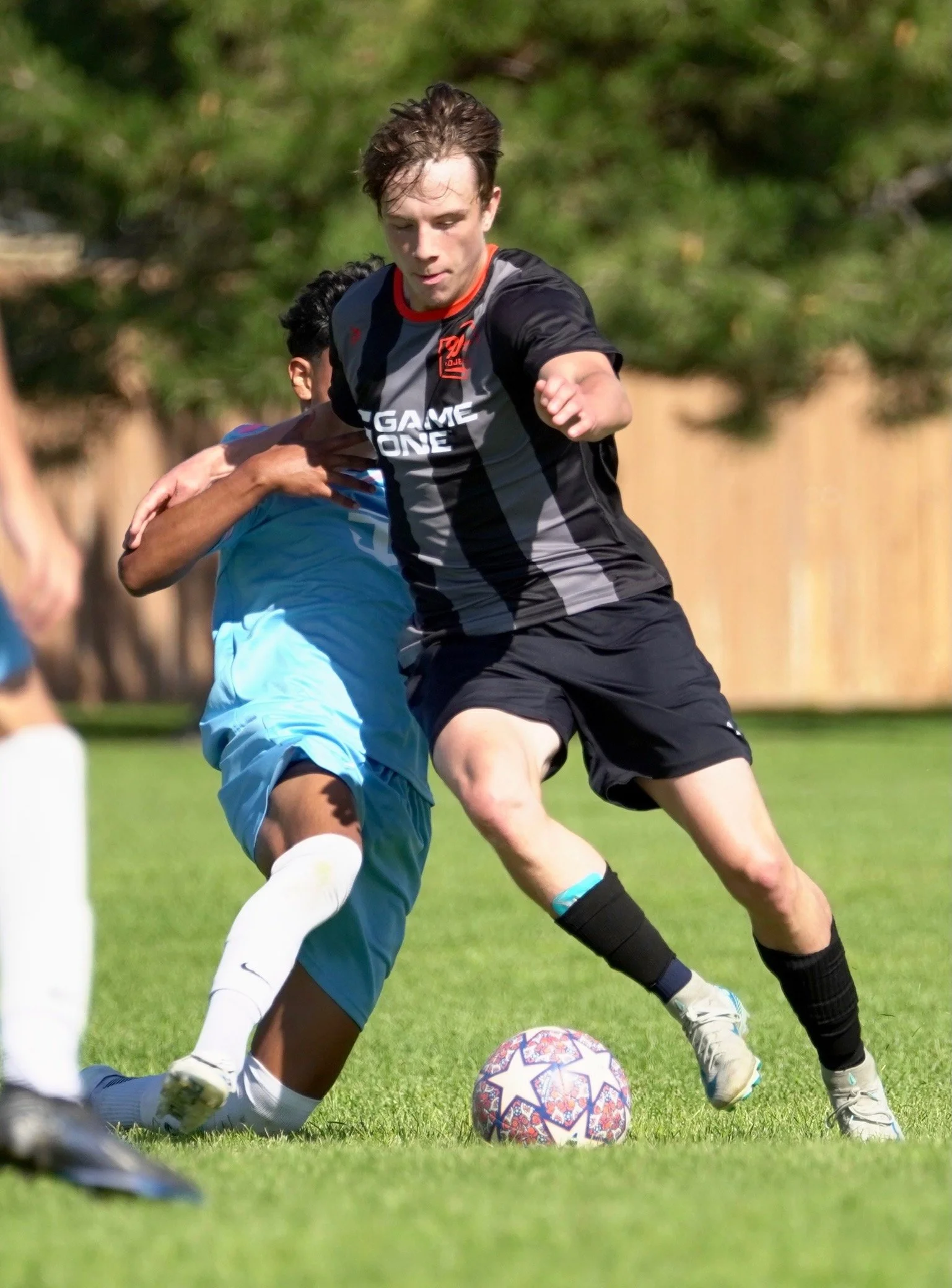 Two male soccer players competing for the ball on a grass field. One player in black and gray uniform, the other in light blue, with trees and a wooden fence in the background.