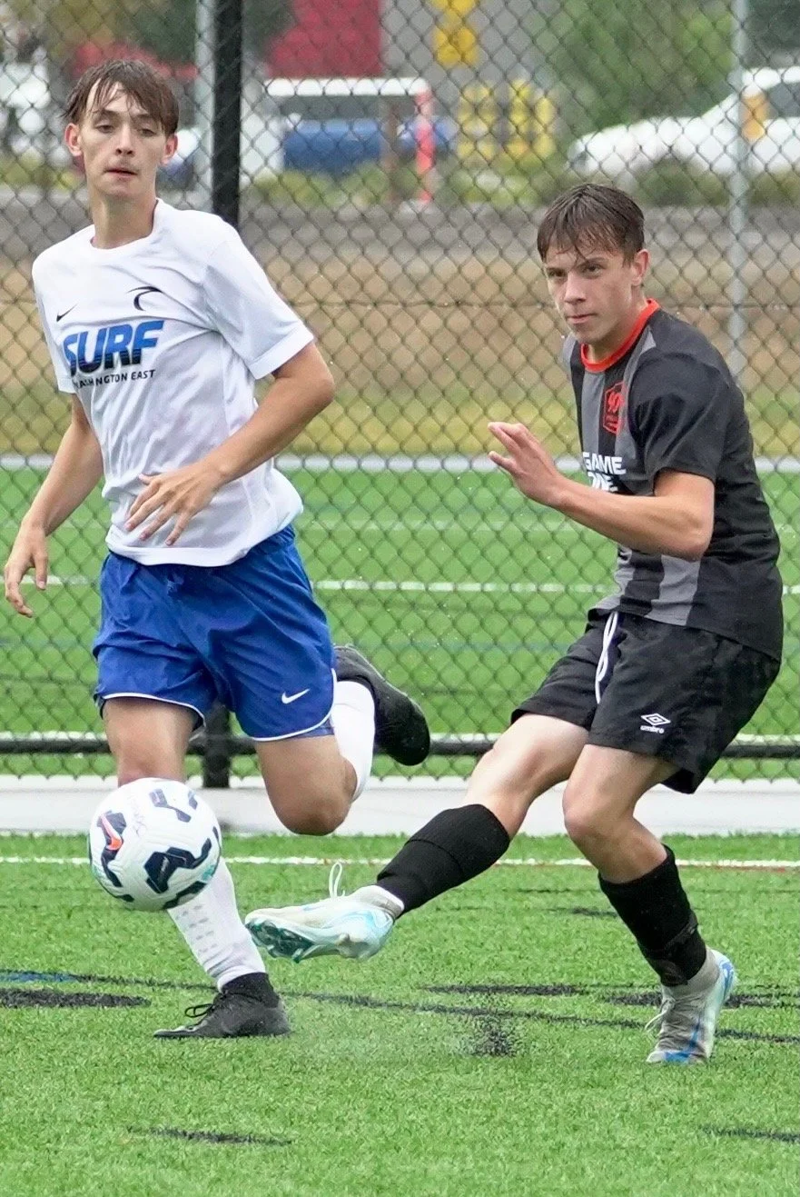 Two teenage boys playing soccer on a grassy field behind a chain-link fence. One boy in a white shirt and blue shorts is kicking the ball, while the other boy in a black and gray uniform is attempting to block or intercept the ball. There is a blurre