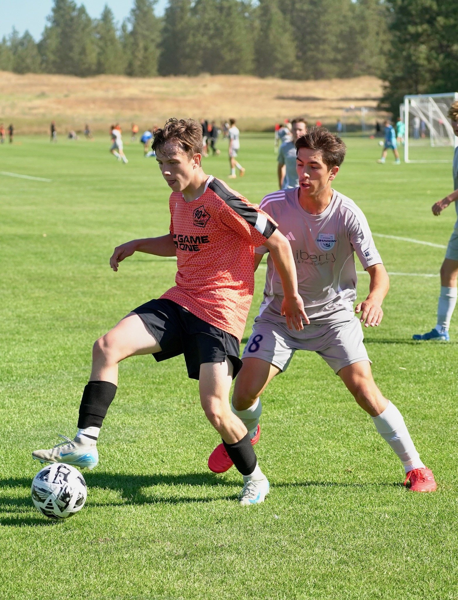 Two boys playing soccer on a grassy field during daytime, with multiple other players in the background.