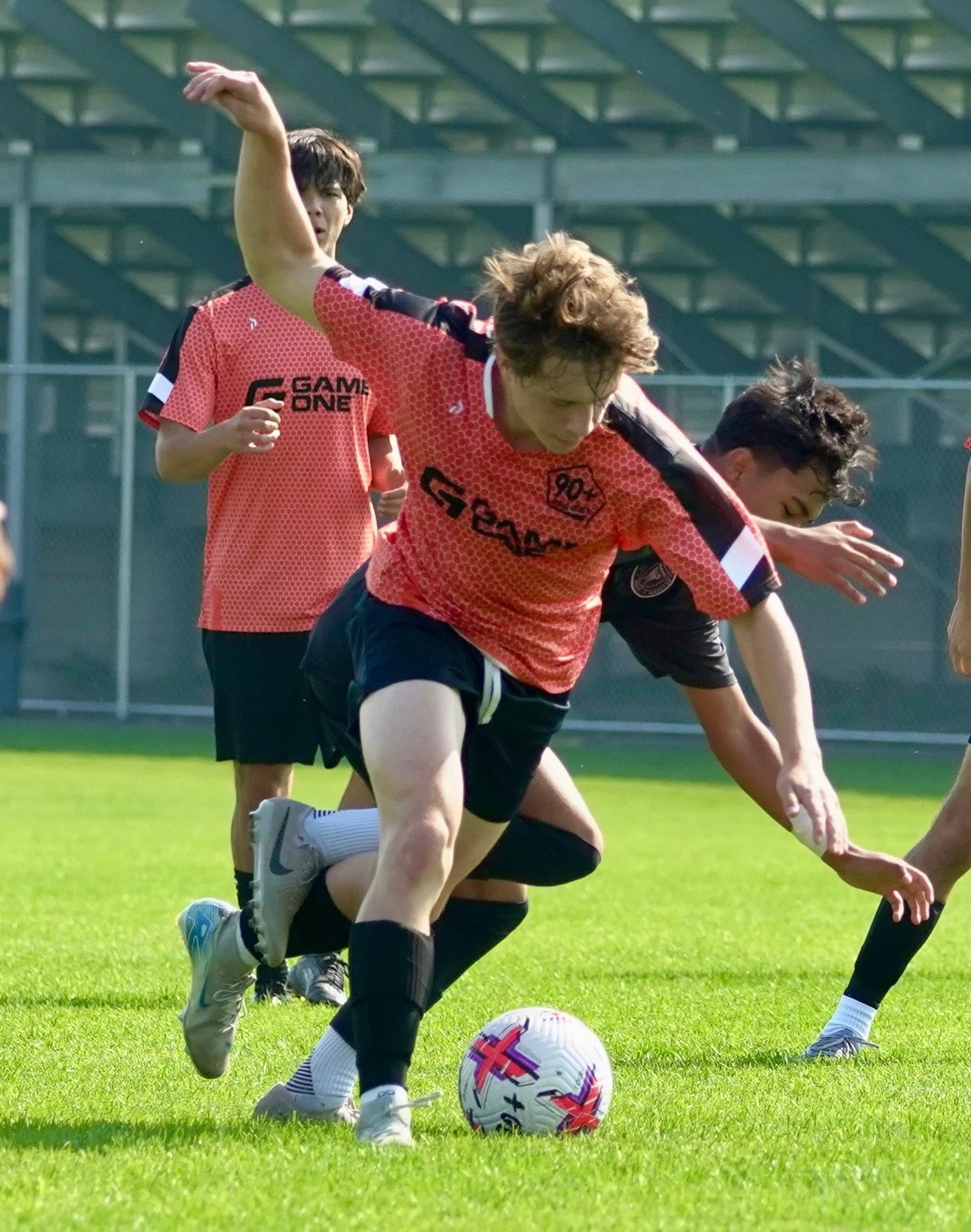 A group of young male soccer players practicing on a grass field. One player is in the foreground, dribbling a soccer ball, wearing a red and black uniform. Others are in the background, also in red and black training gear.