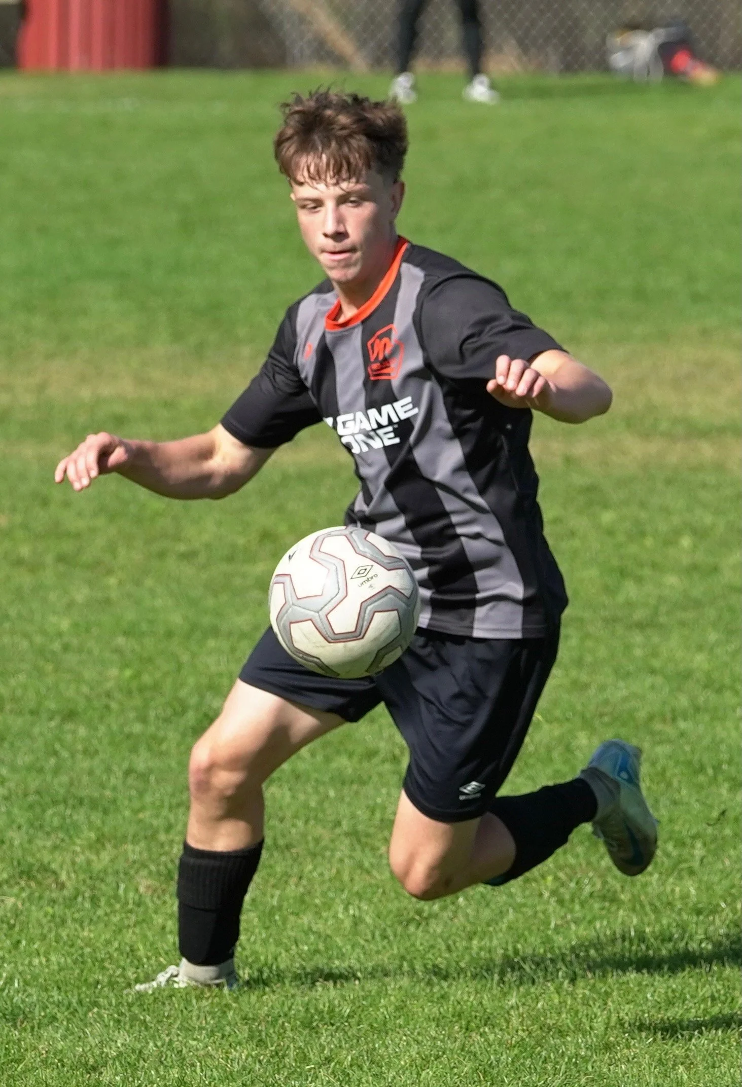 A young male soccer player in a black and gray uniform is kicking a soccer ball on a green field.