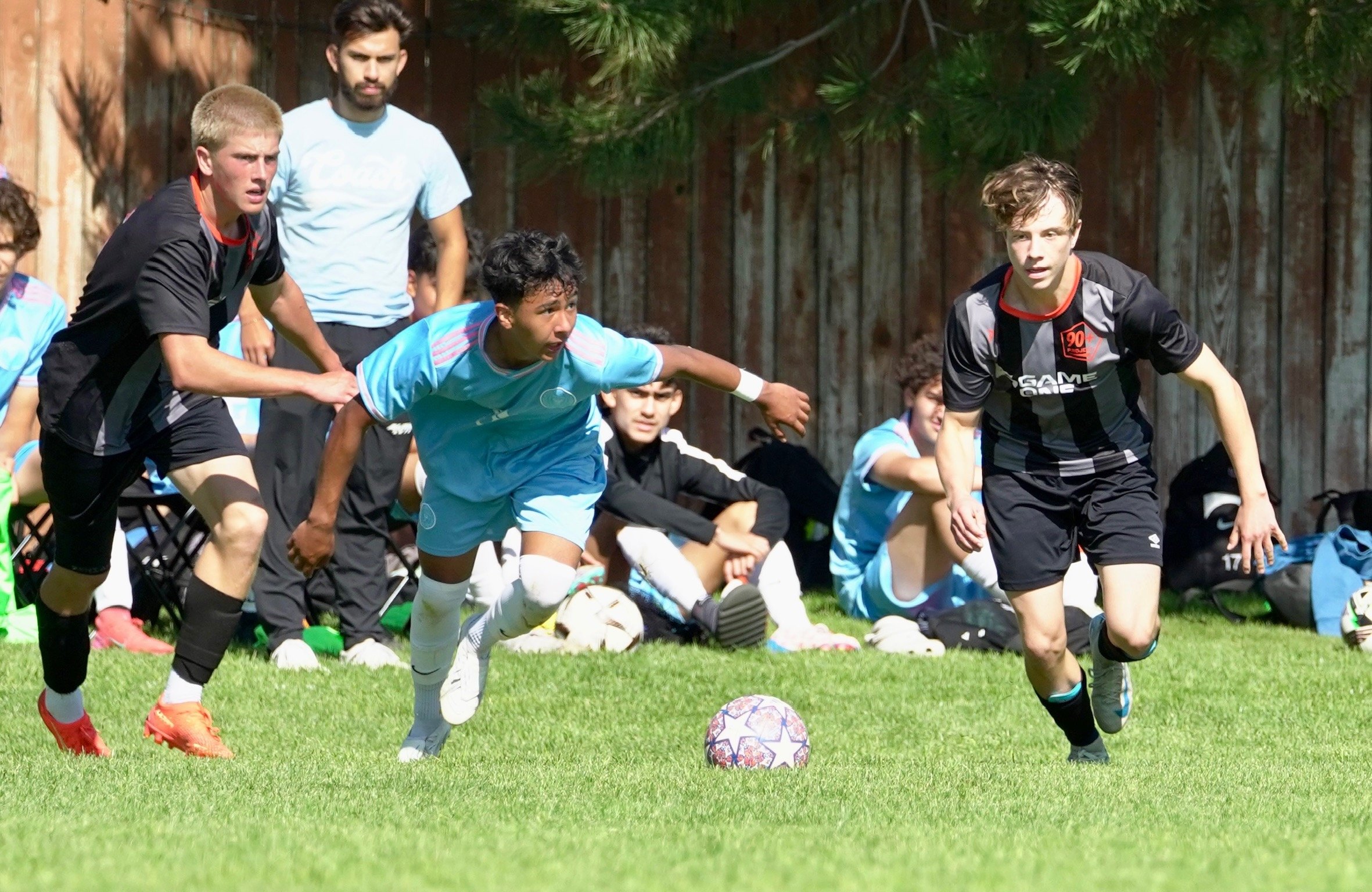 Two young soccer players racing for the ball during a game, with spectators sitting on the bench in the background.