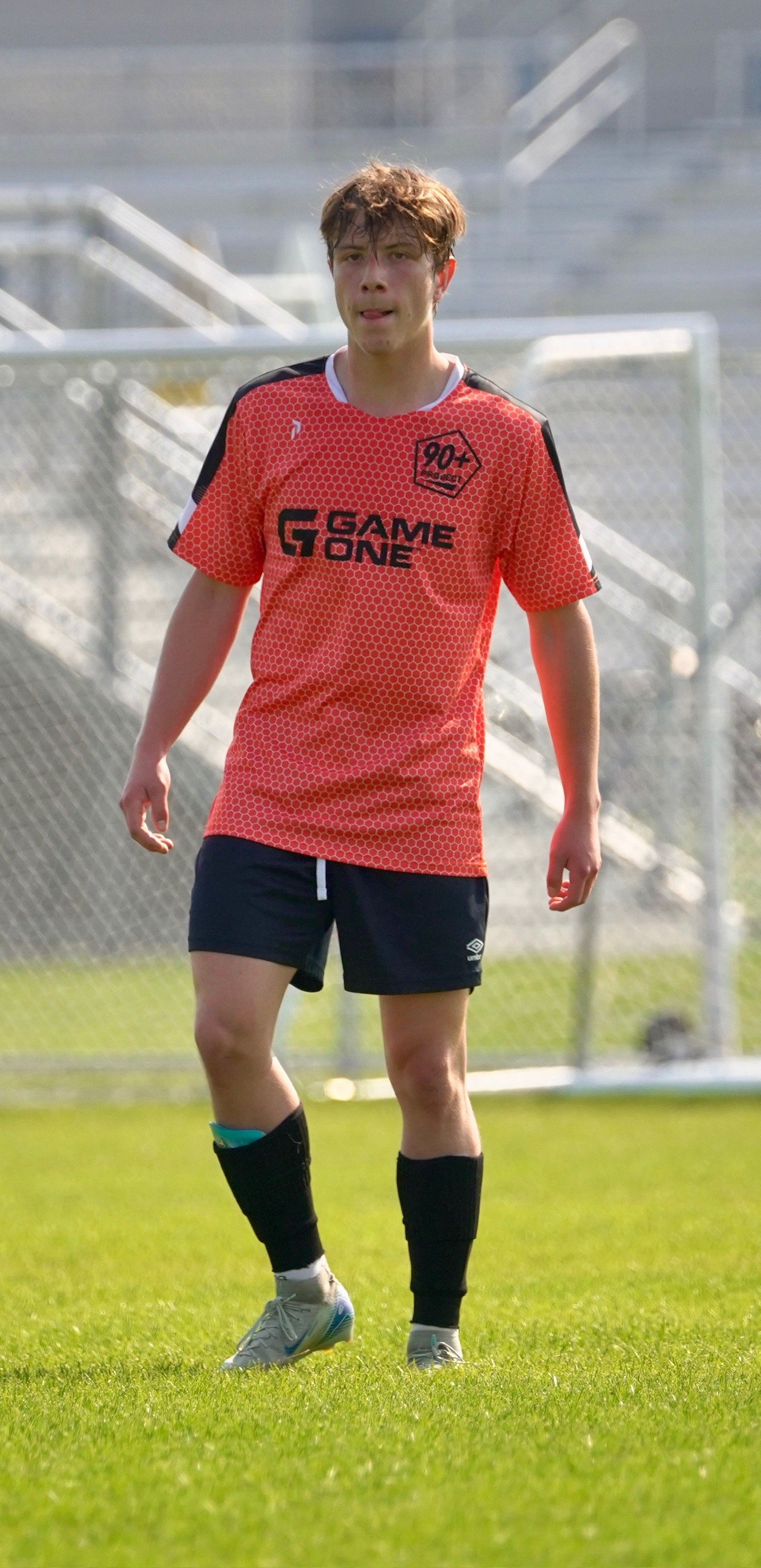 A young man playing soccer on a field, wearing a red jersey with black accents, black shorts, black socks, and gray soccer cleats.