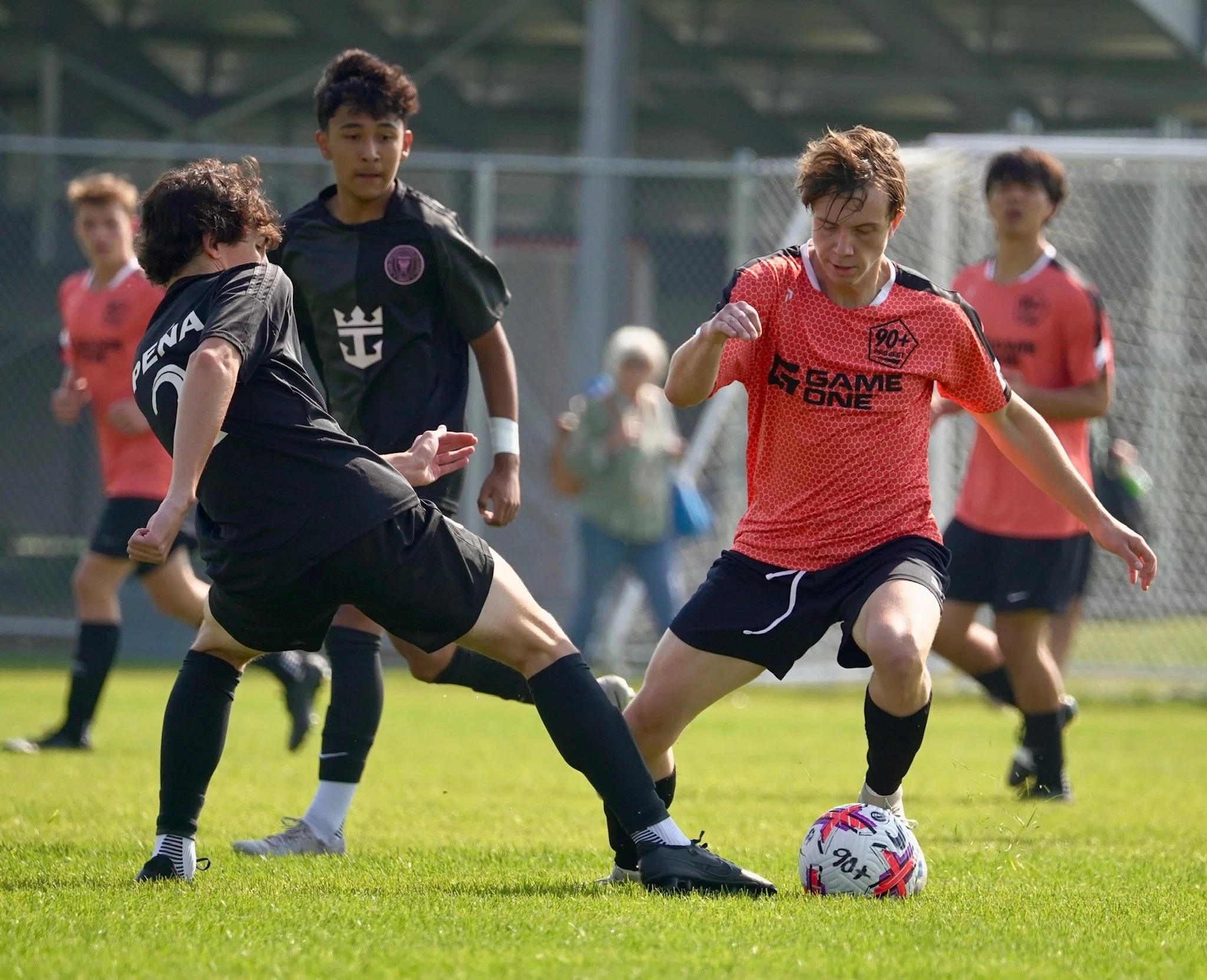 Young male soccer players engaged in a match on a grassy field, with one player kicking the ball and others watching, set outdoors with a metal fence and spectators in the background.