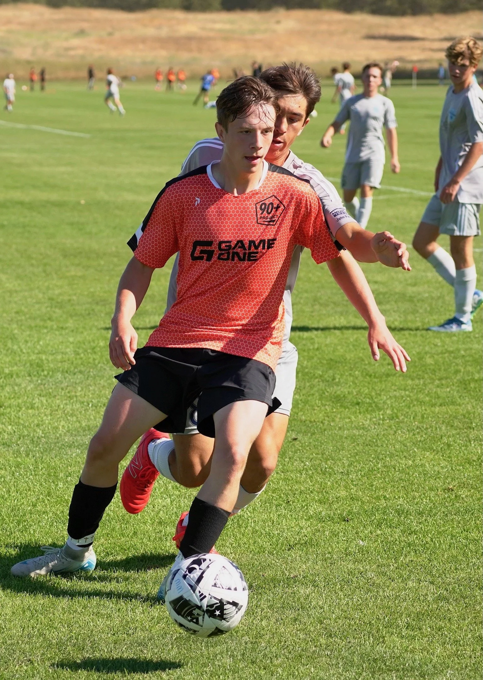 Two young male soccer players compete for control of a soccer ball on a grassy field during a match. The player in front wears a red-orange jersey and black shorts, while the player behind wears a gray jersey. Several other players are visible in the