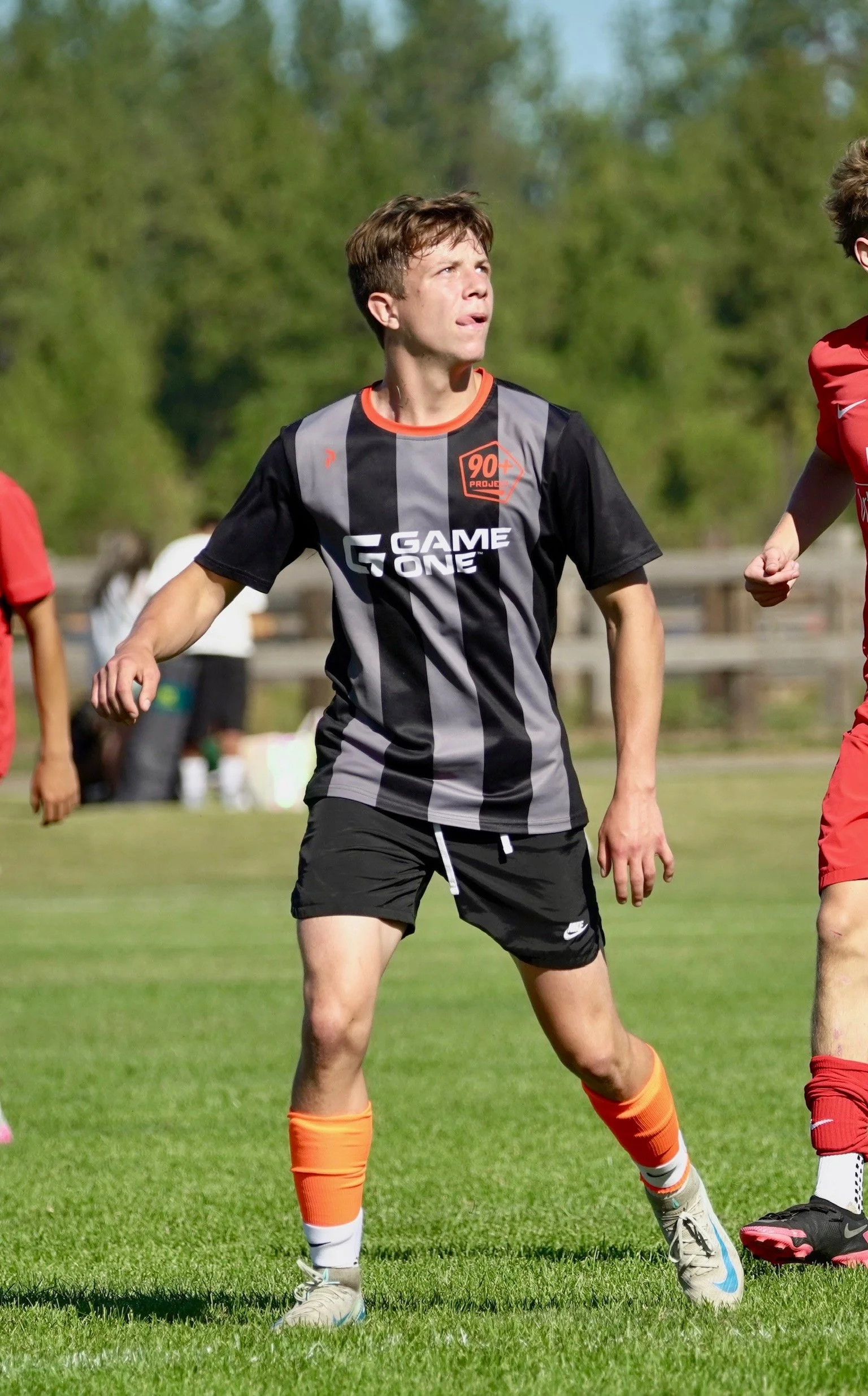 A young male soccer player in a black and gray striped jersey with orange details, black shorts, and orange socks on a grassy field, looking to the left during a game.