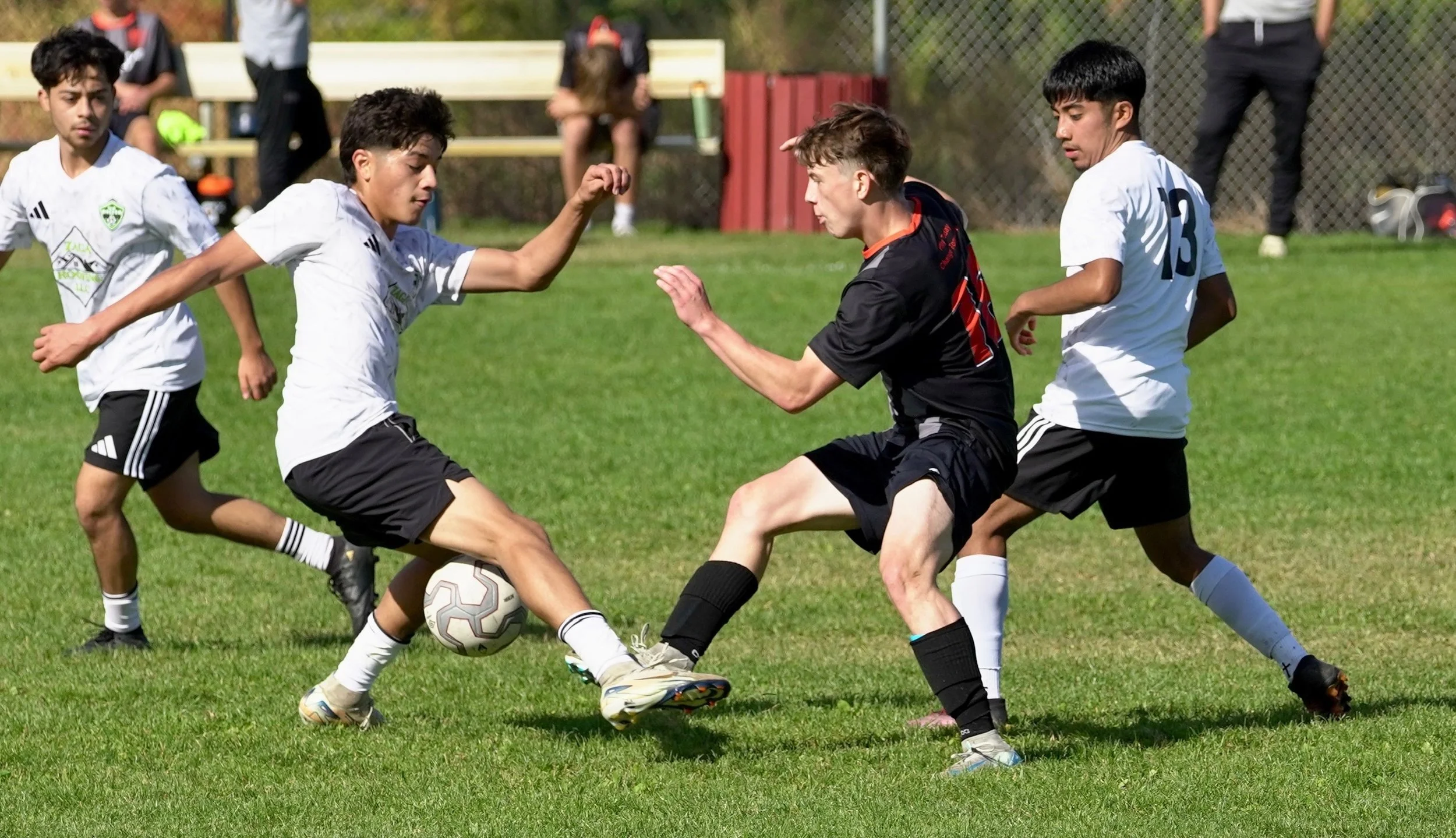 Four young male soccer players are contesting for the ball on a grassy field during a match, with some spectators and a fence in the background.