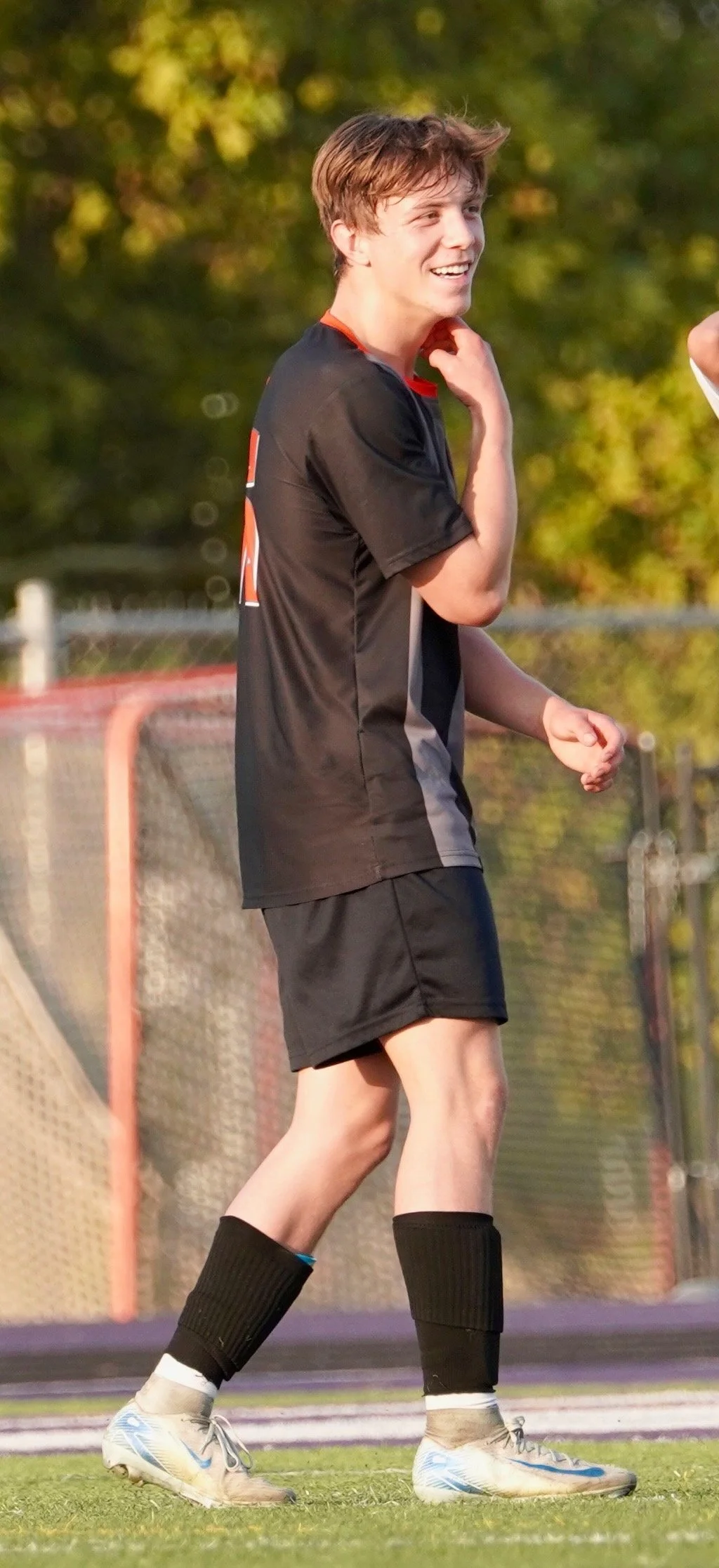 A young man in a black sports uniform with tall black socks and white cleats, standing on a sports field, smiling and adjusting his collar.