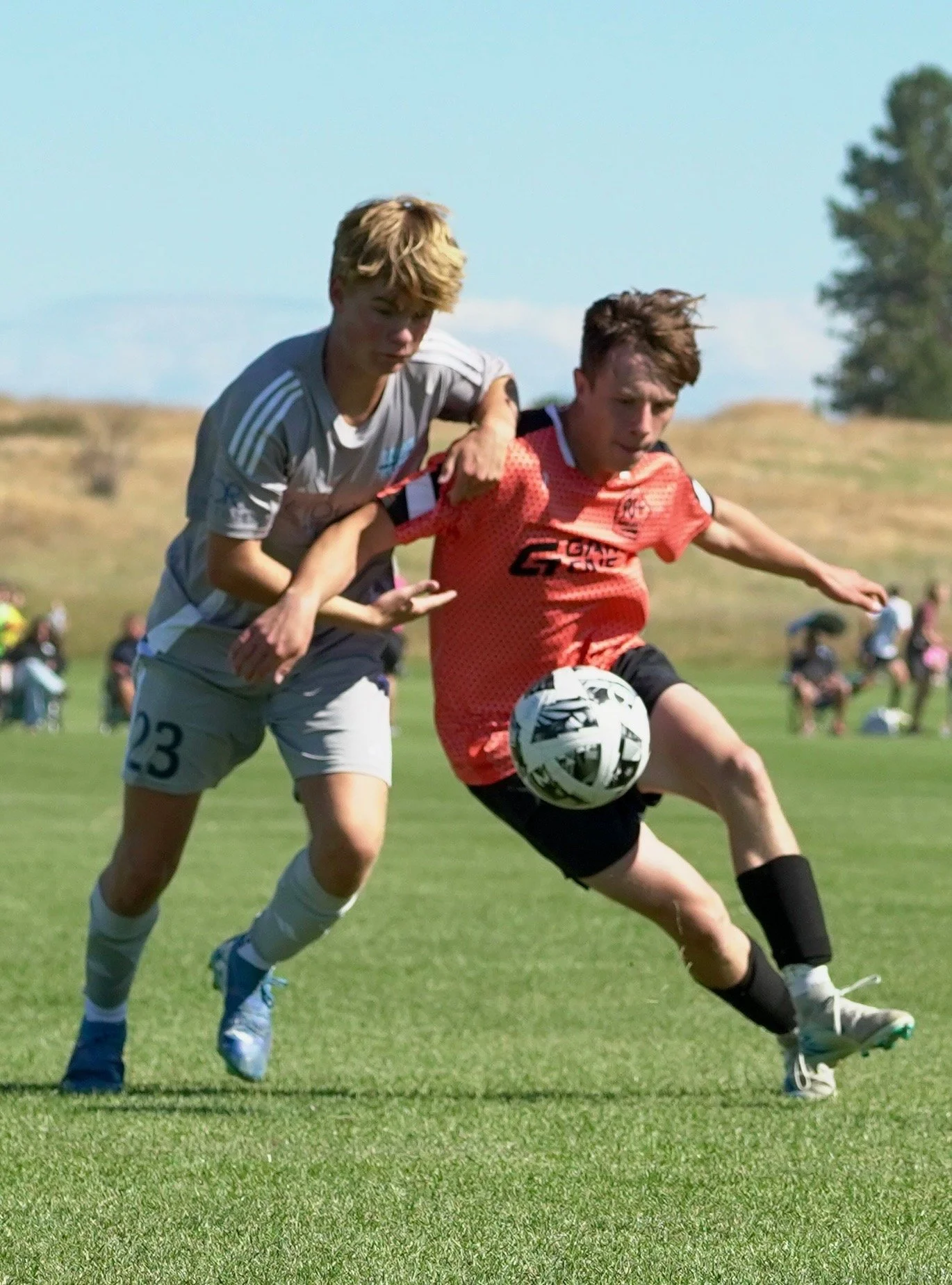 Two boys playing soccer outdoors, with one in a red jersey kicking the soccer ball and the other in a gray jersey trying to block him, on a grassy field under a blue sky.