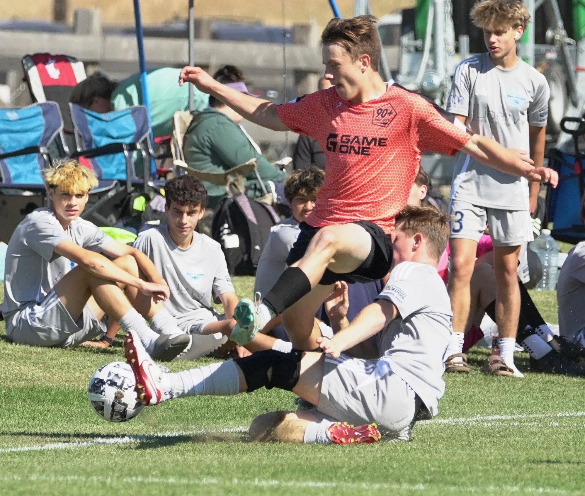 A youth soccer match showing a player in white covering a soccer ball with his body while a player in red jumps and tackles him on the field as other players and spectators watch nearby.