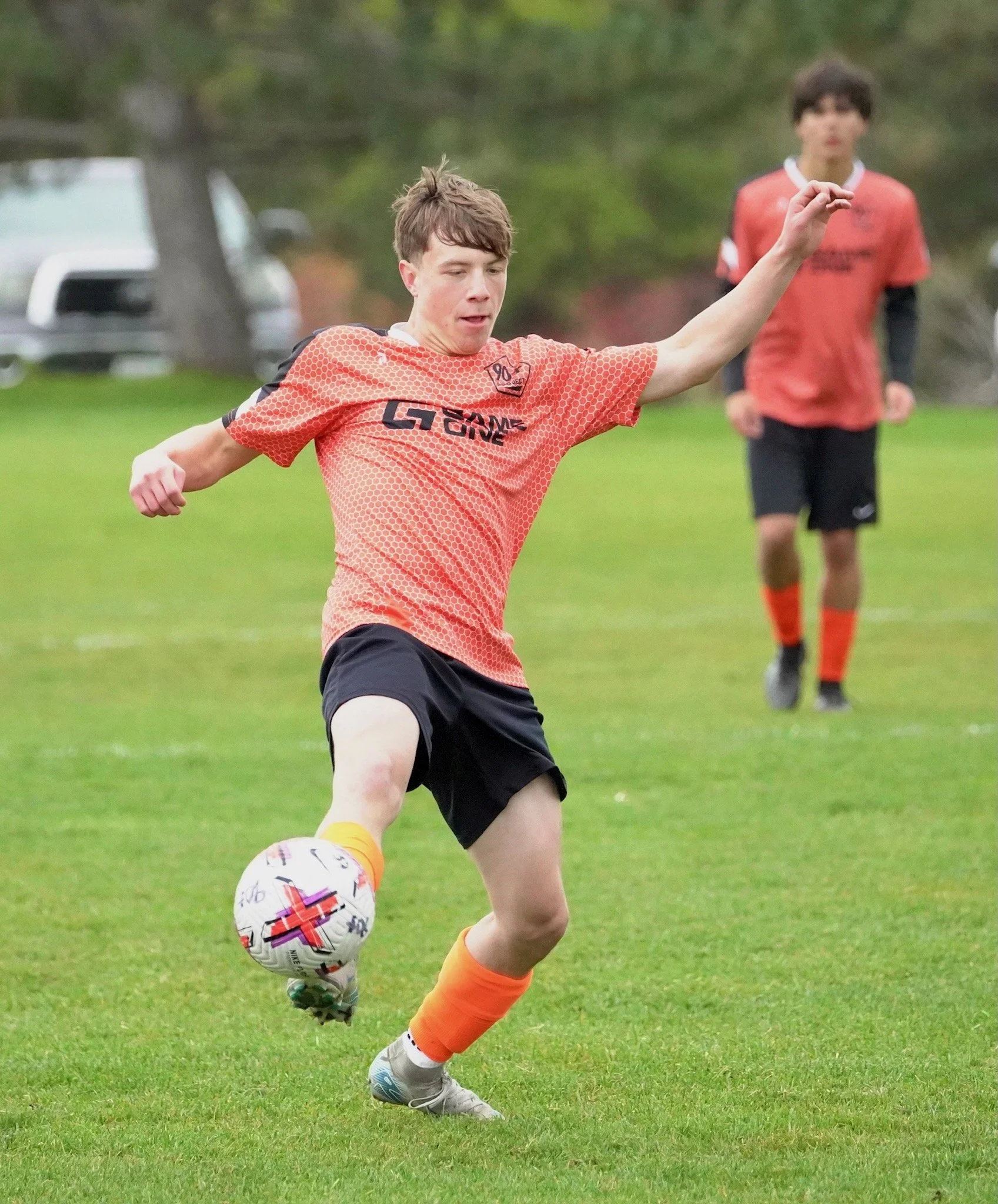A young man wearing an orange sports jersey and black shorts is kicking a soccer ball on a grassy field. Another young man in similar attire is in the background.