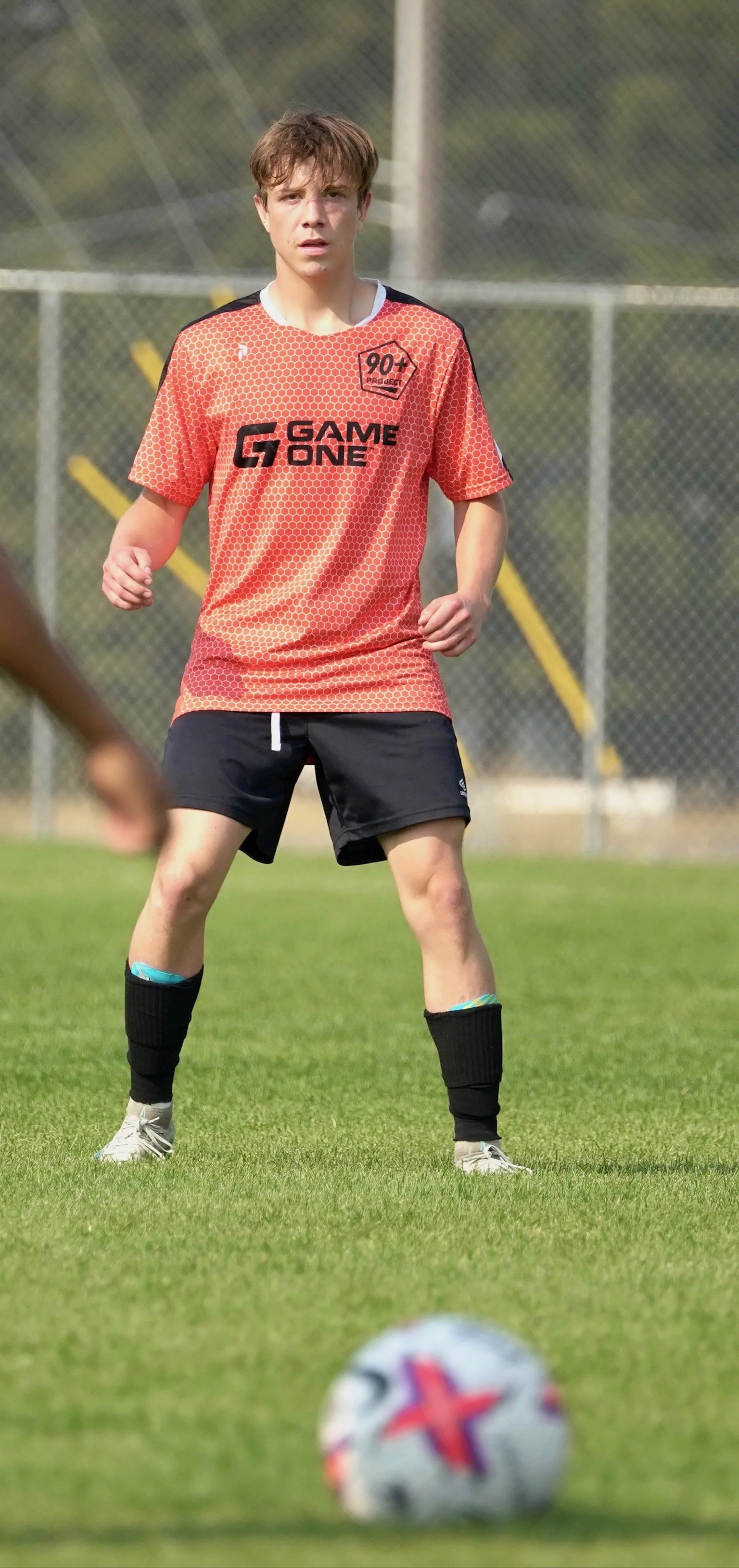 A young man playing soccer on a field, wearing a red and black sports jersey, black shorts, and black socks, preparing to kick a soccer ball.