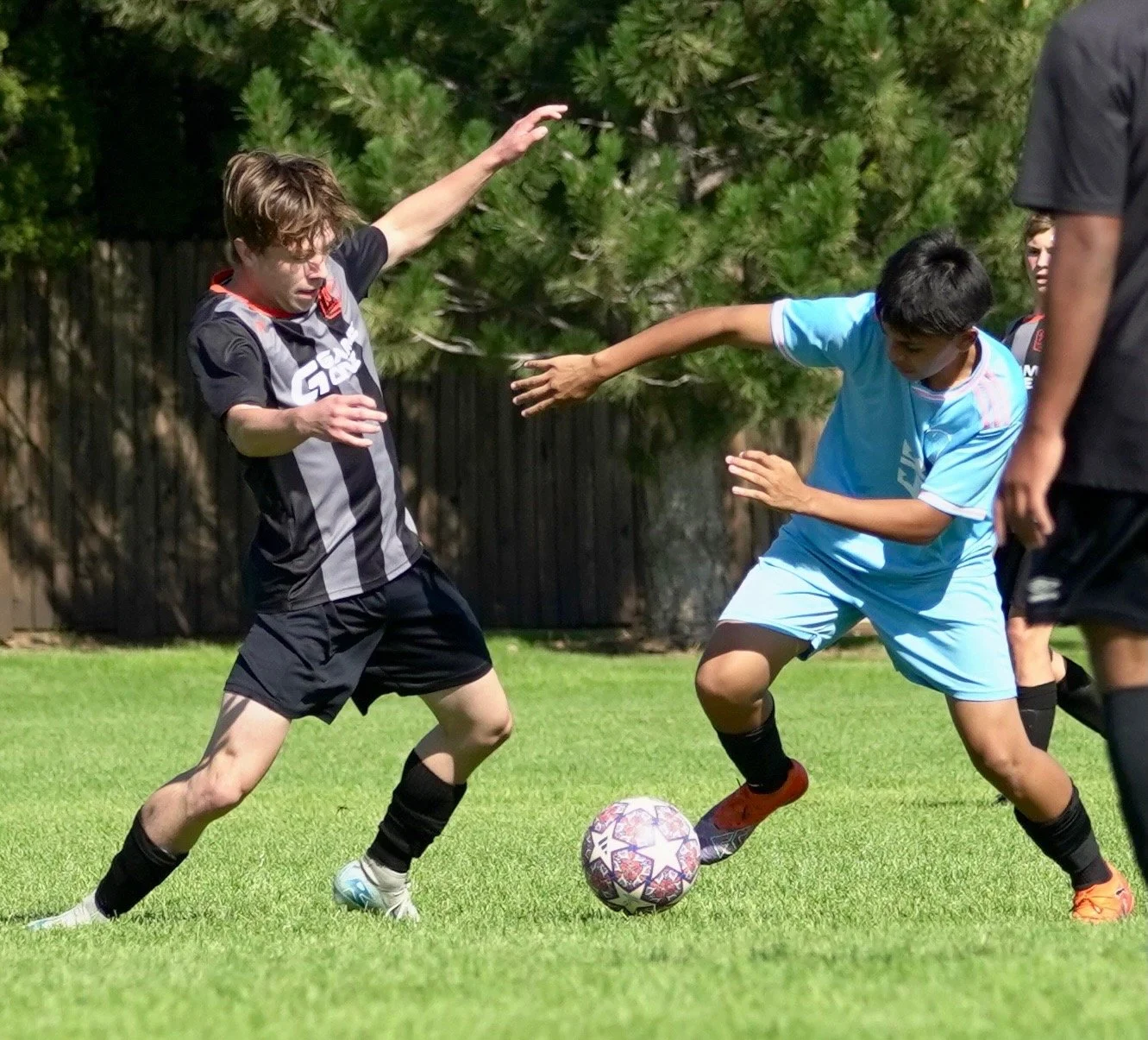Two young boys playing soccer outdoors on a green field, competing for the ball with a third player partially visible on the right.
