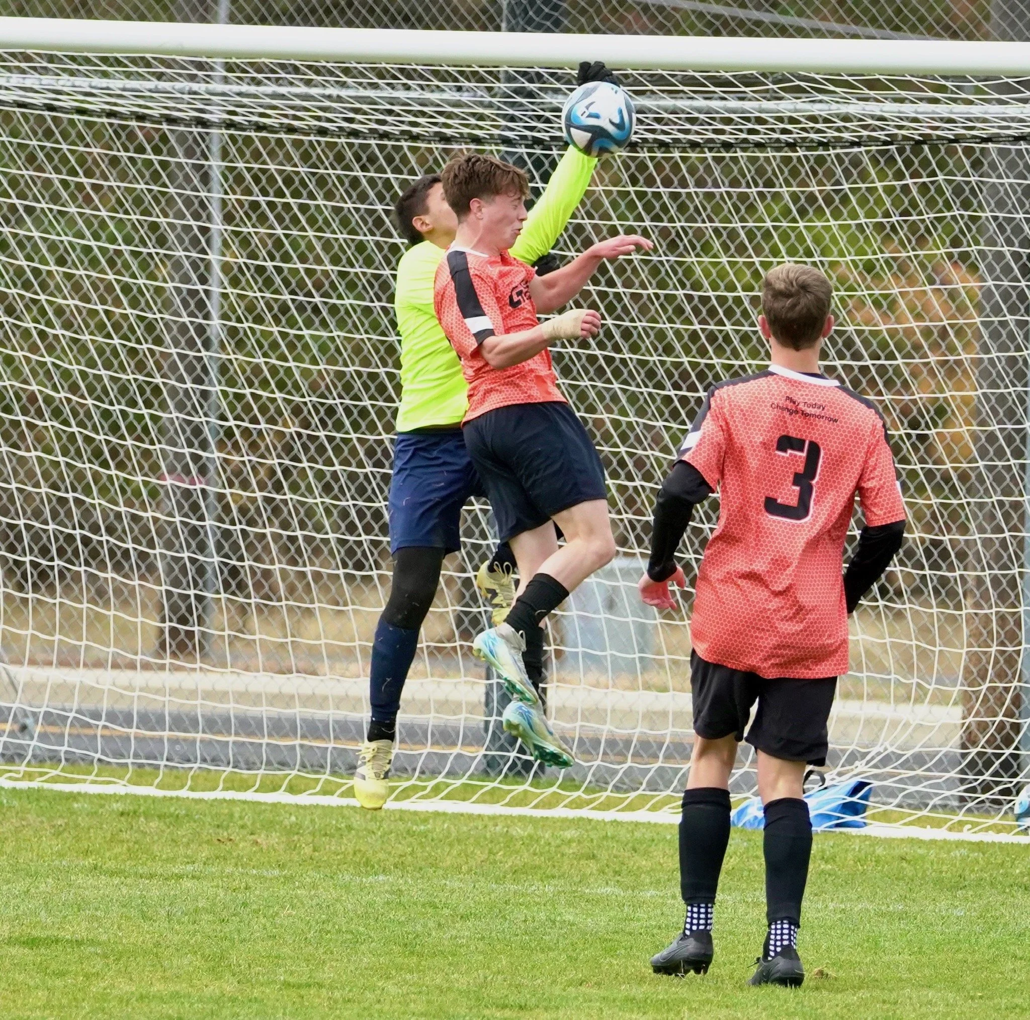 A youth soccer game showing a goalie in a bright green jersey catching a ball near the goal, with a player in an orange jersey jumping to block, and another player in an orange jersey with the number 3 watching from behind.