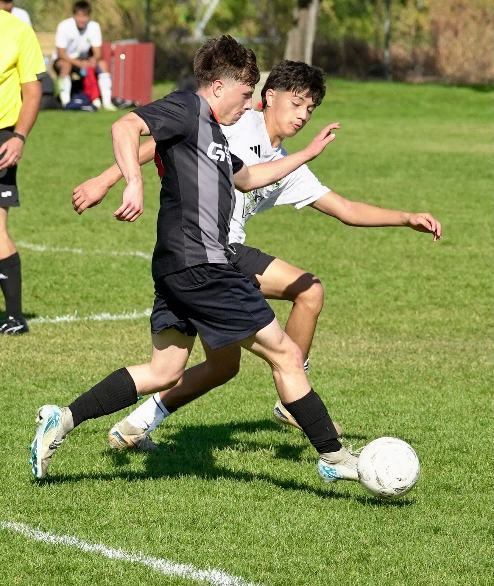 Two young soccer players compete for the ball on a grassy field, one in a black uniform and the other in a white uniform, with a referee and other players in the background.