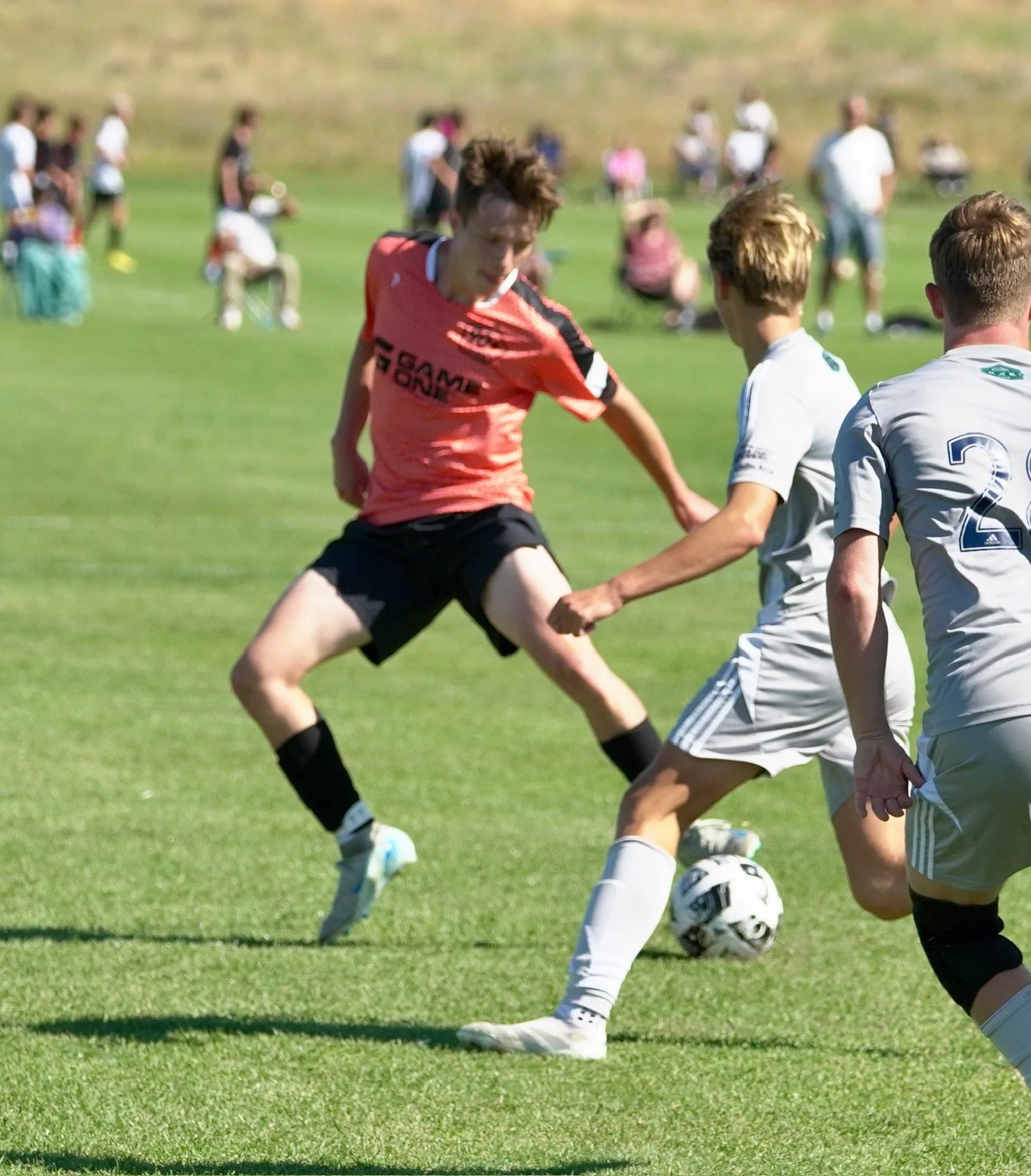 Three young boys playing soccer on a grassy field, with one in a pink shirt and two in white uniforms, actively competing for the ball, with spectators watching in the background.