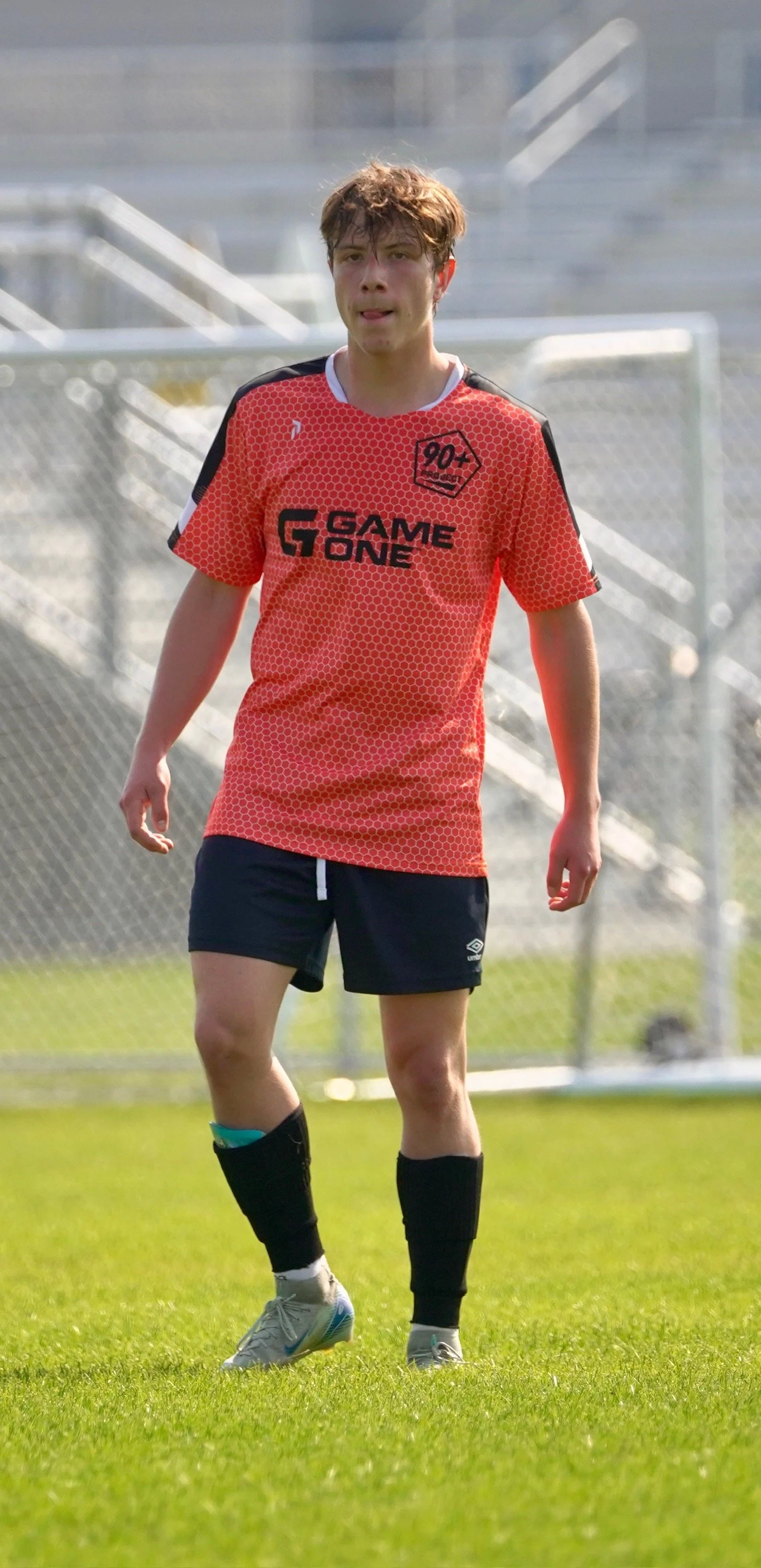 Young male soccer player in red jersey with black shorts and black socks on a soccer field.