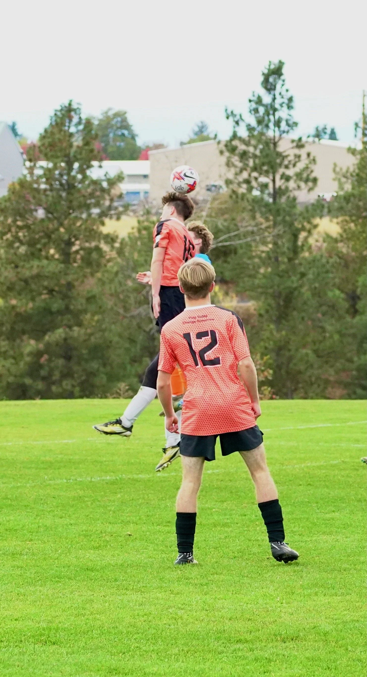 Young male soccer players in orange jerseys and black shorts playing on a grassy field, one jumping to head the ball, others watching, with trees and buildings in the background.