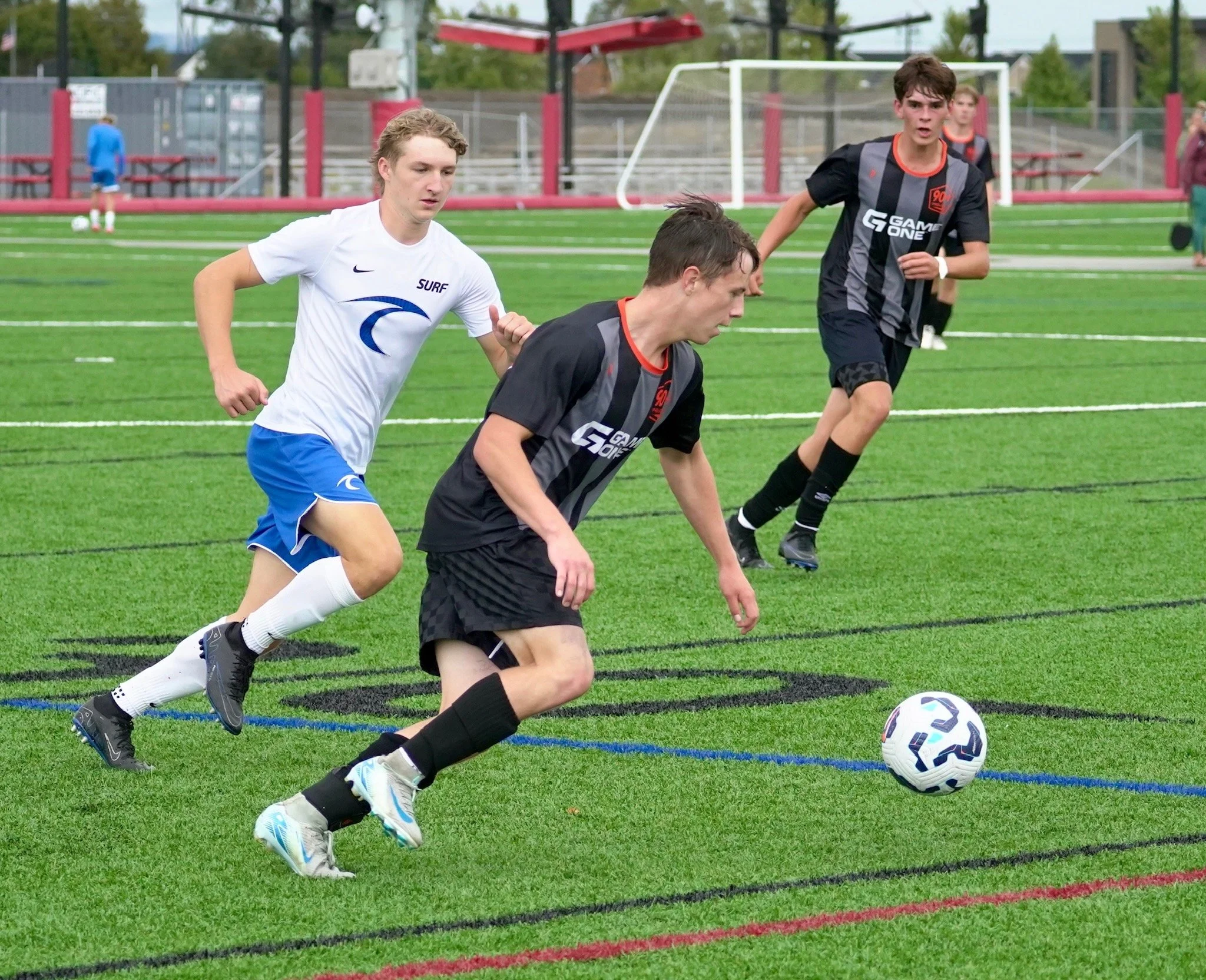 Three young men playing soccer on a green field, with one player in black chasing the ball while two players in white and blue follow. In the background, a soccer goal and other people are visible.
