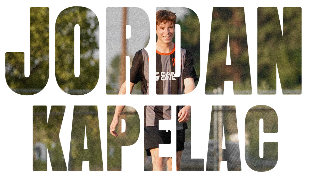 A young boy with curly brown hair wearing a black and gray soccer uniform with orange accents and a sponsor logo, walking outdoors on a sunny day with trees and a chain-link fence in the background.