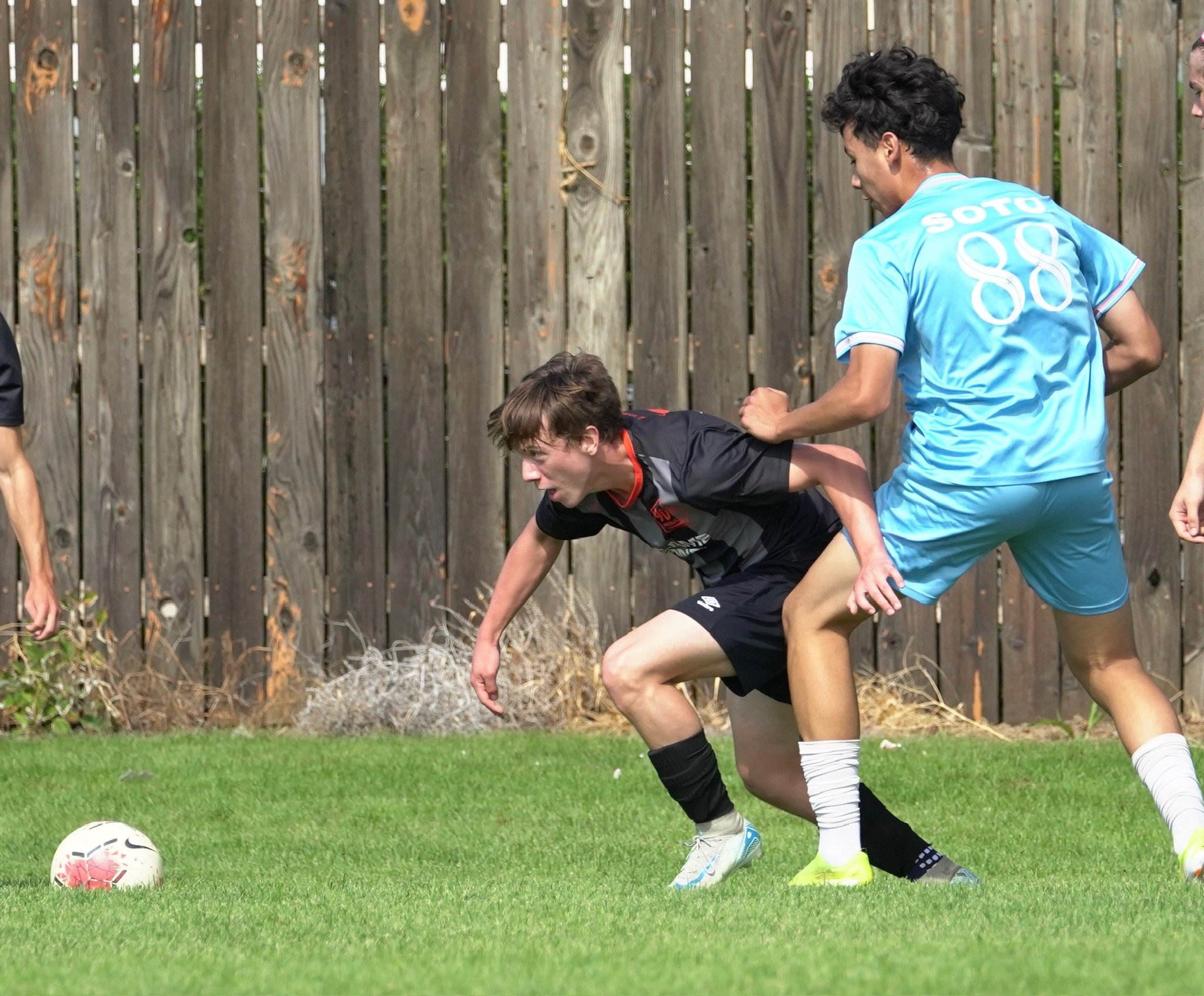 Two young men playing soccer on a grassy field near a wooden fence. One in a black jersey is bending down to control the ball, while the other in a blue jersey with the number 88 is defending.