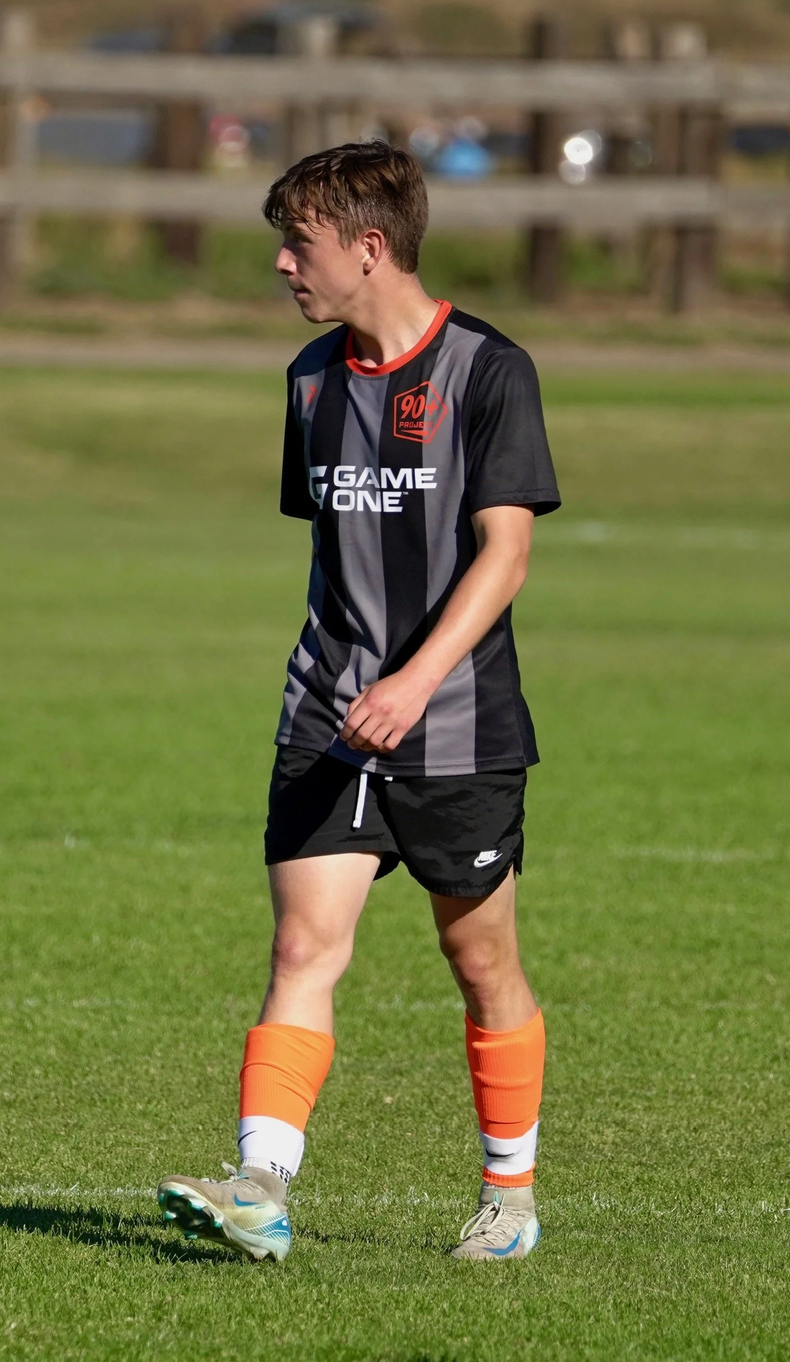 A young male soccer player in a black and gray jersey with the text 'Game One' and a logo, black shorts, orange socks, and gray cleats standing on a grassy field.
