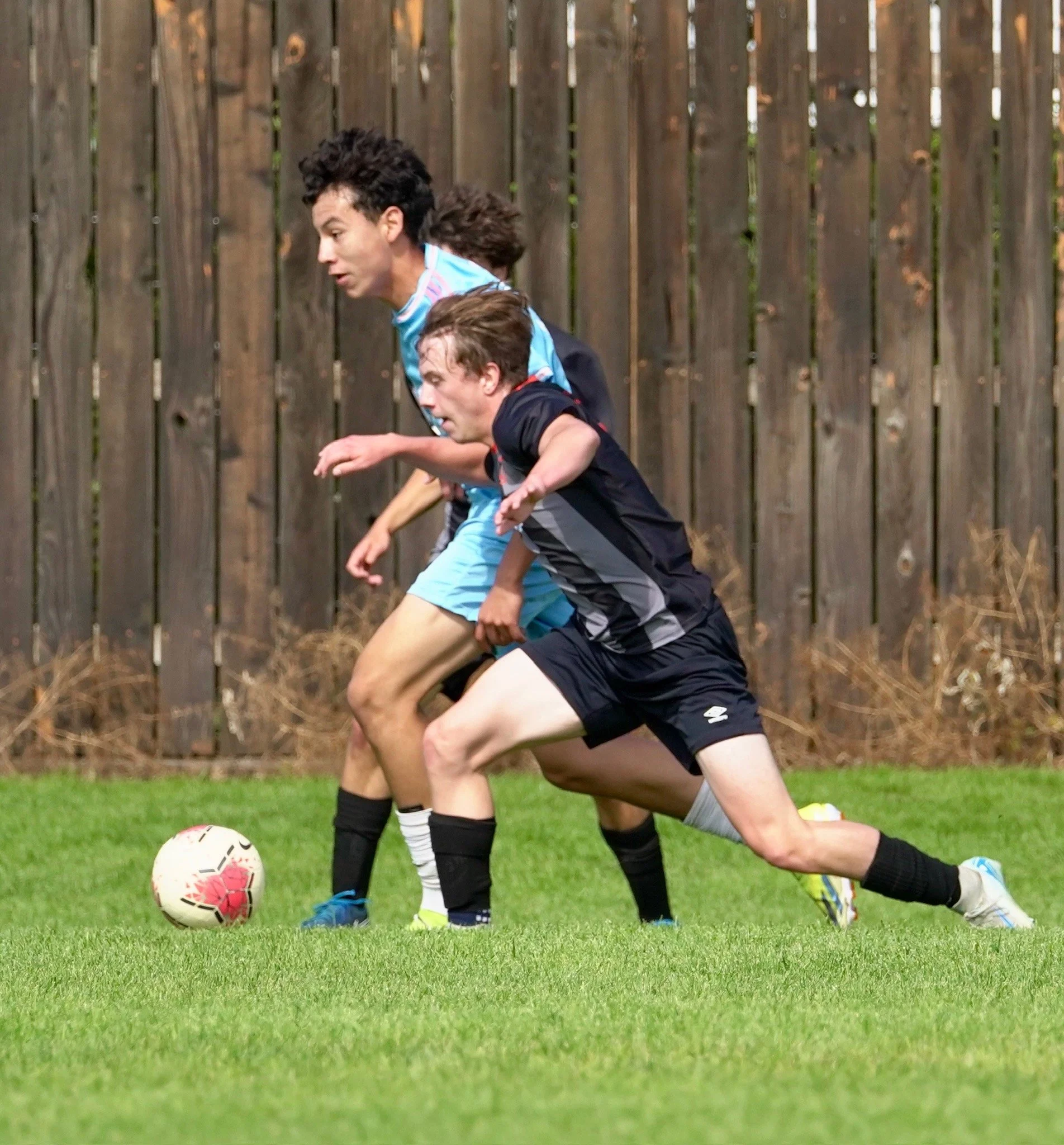 Three young men playing soccer outdoors, focusing on the ball, with a wooden fence in the background.