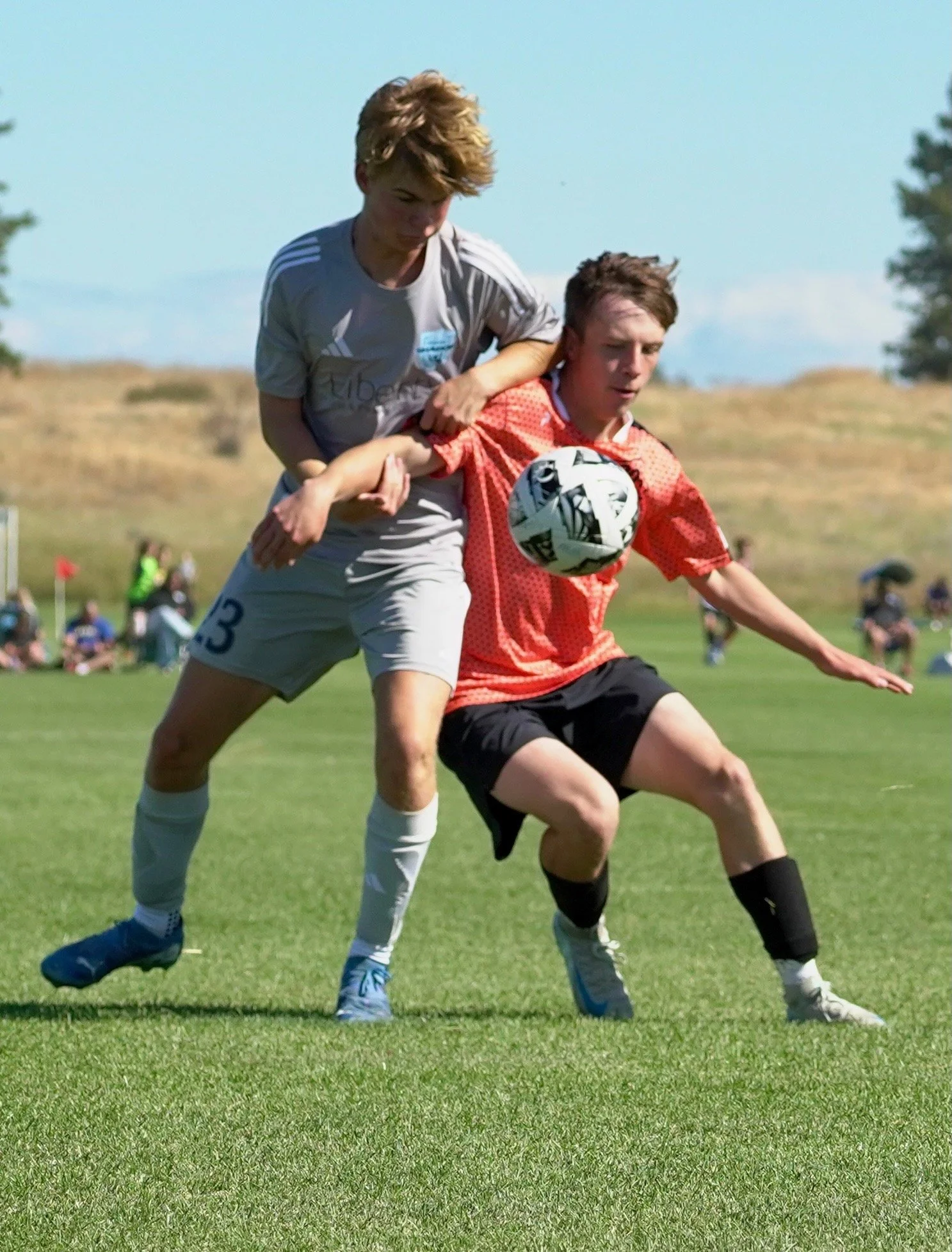 Two young boys playing soccer outdoors, one in a gray uniform and the other in a red jersey, competing for the ball during a match on a green field with a scenic landscape in the background.
